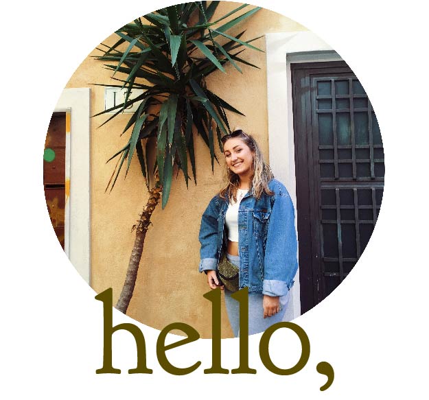 white blonde female artist, posing next to a palm tree, on a yellow painted wall on the left hand side. on the right side there is a gothic dark brown door. the female is wearing a denim jacket, white vest top, an olive sparkling bum bag, grey leggings and has long blonde flowing hair.