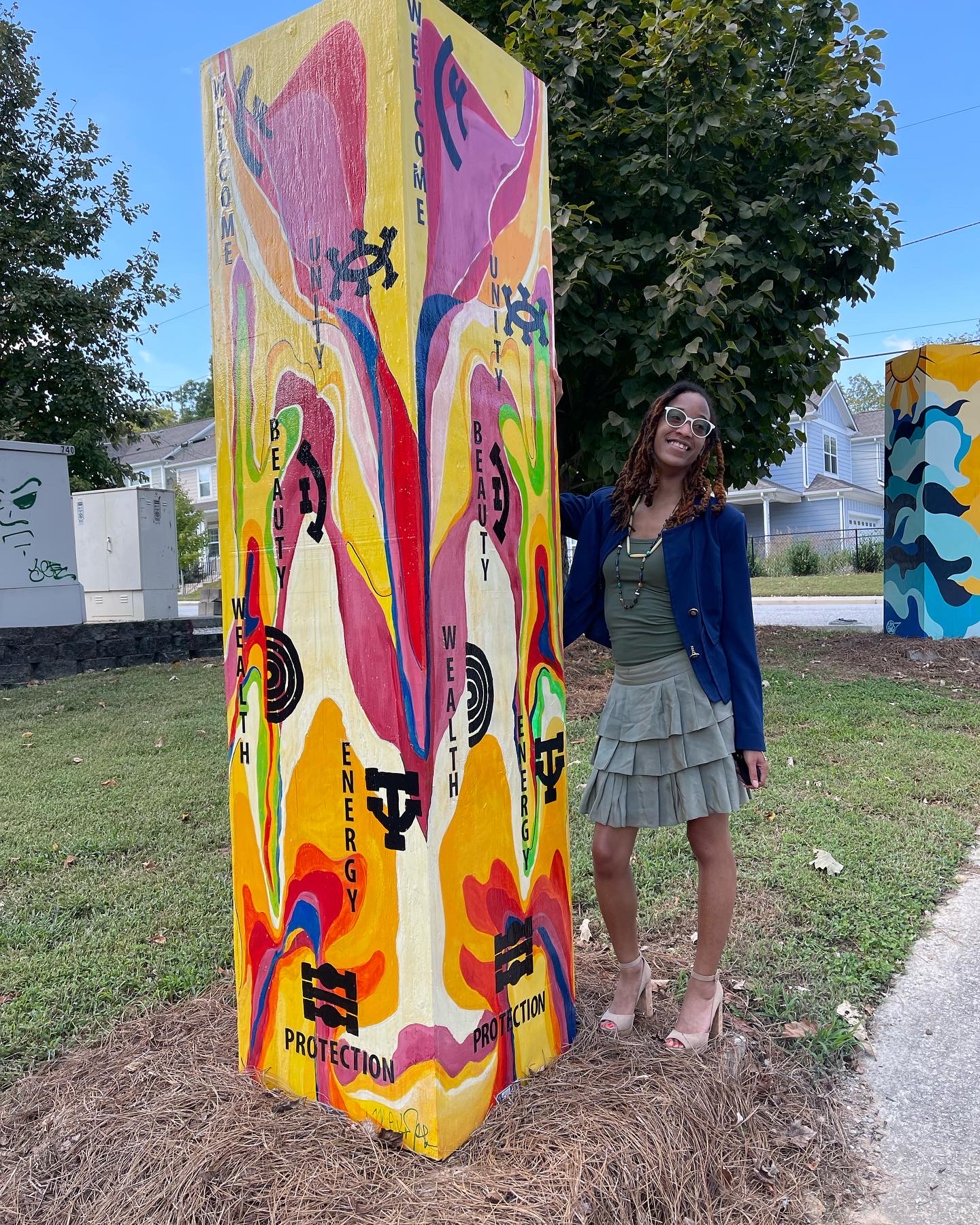 Mia Anika stands next to wooden pillar painted orange, red, blue, green with black labels that read, Welcome and Unity.