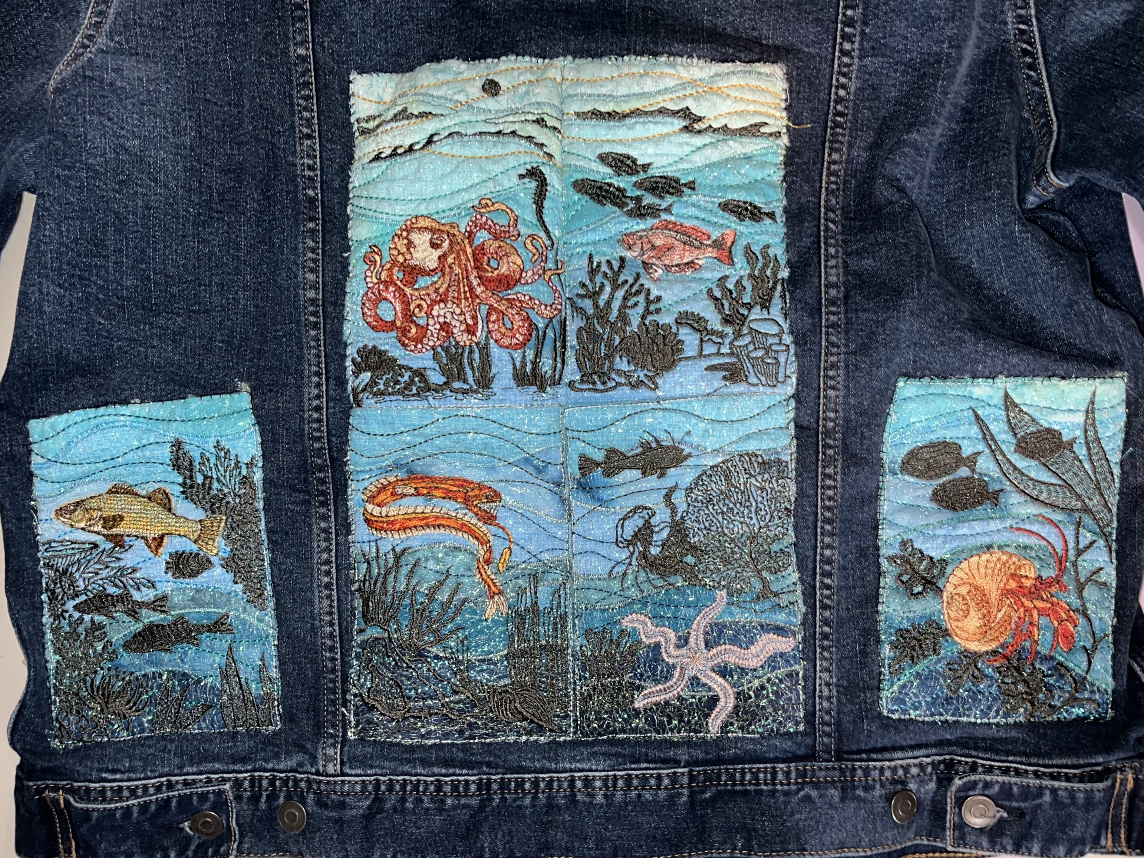 Blue jean jacket with six quilted embroidery panels of sea creatures, including an octopus, rockfish, dragonfish, brittle star, hermit crab, and another green fish.