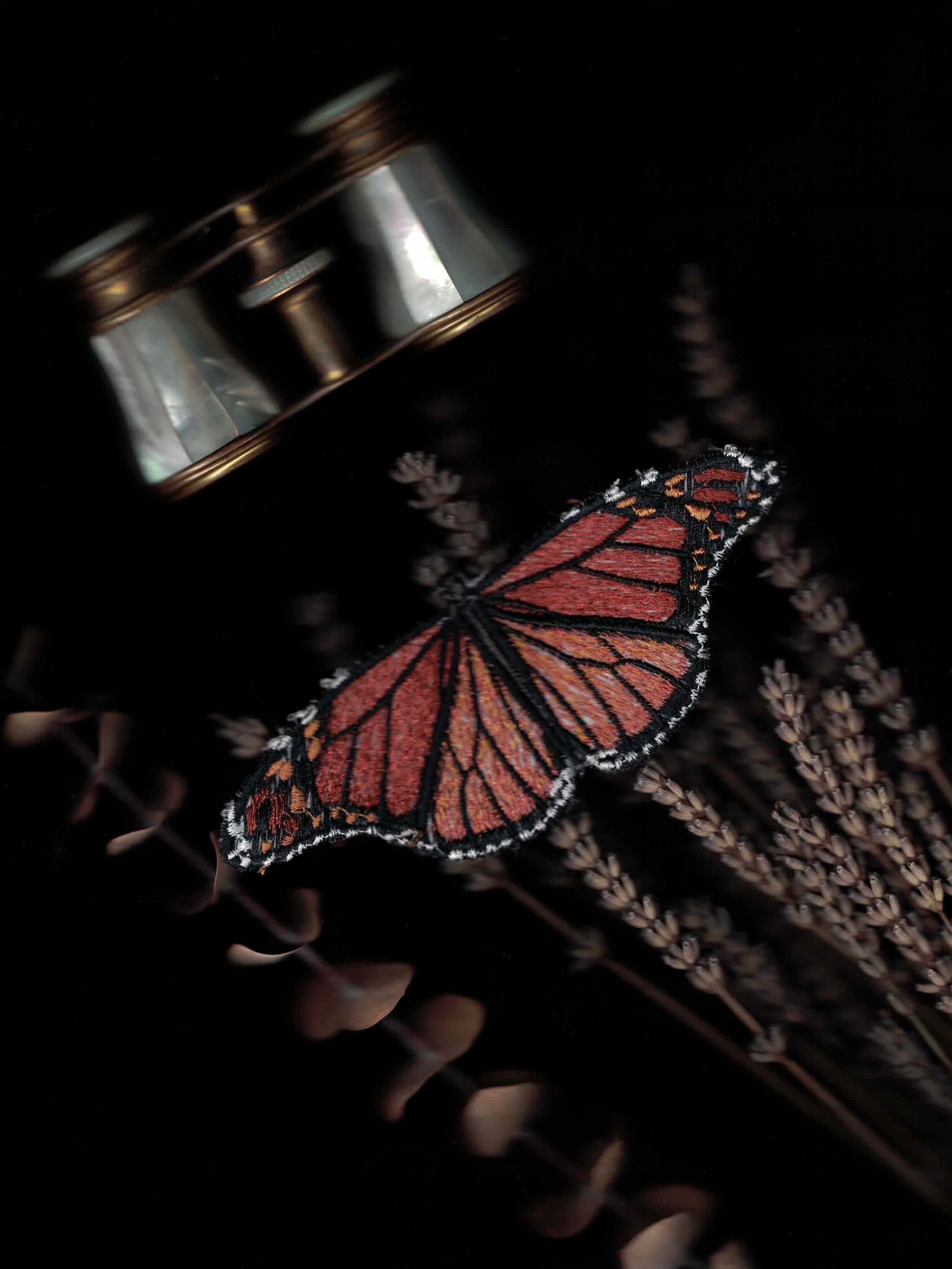 Embroidered monarch butterfly perched on lavender being watched by a pair of mother of pearl opera glasses.