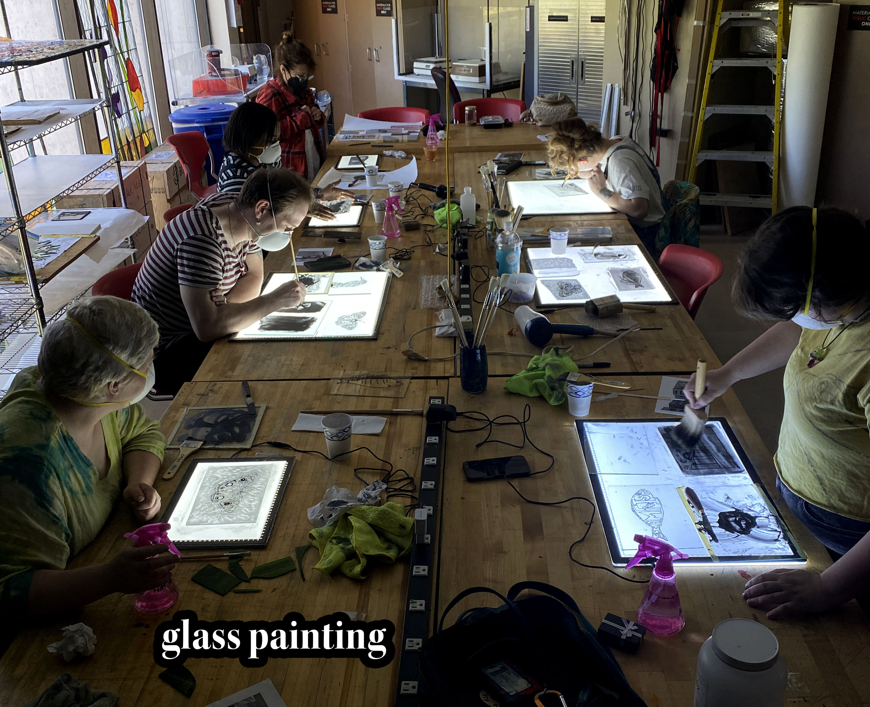 students hovering over illuminated LED light pads, focusing on their glass paintings in front of them