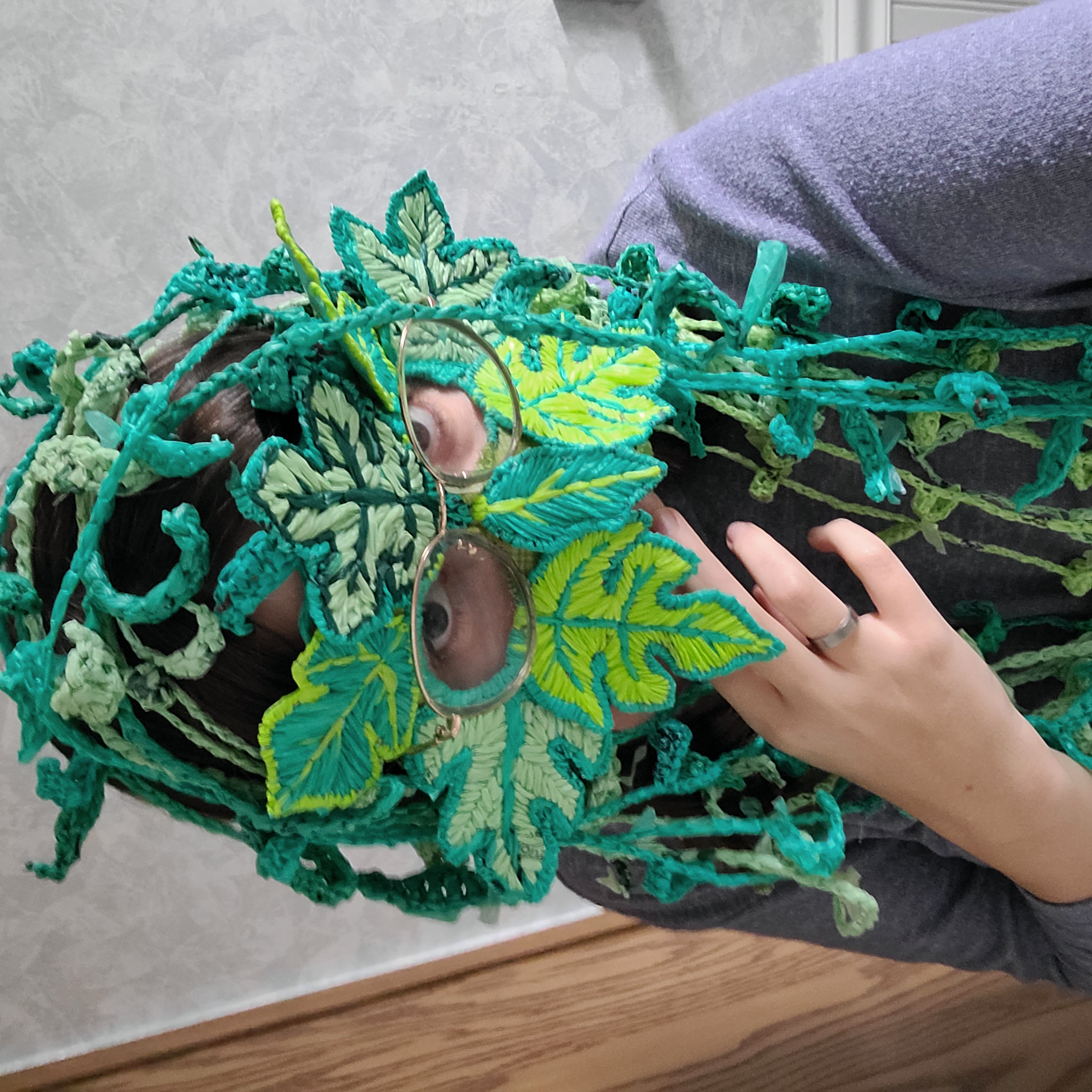 Domenica close up wearing a "green man" mask made of leaves that are constructed of embroidered plastic and wearing crocheted plastic willow leaf garland on her head