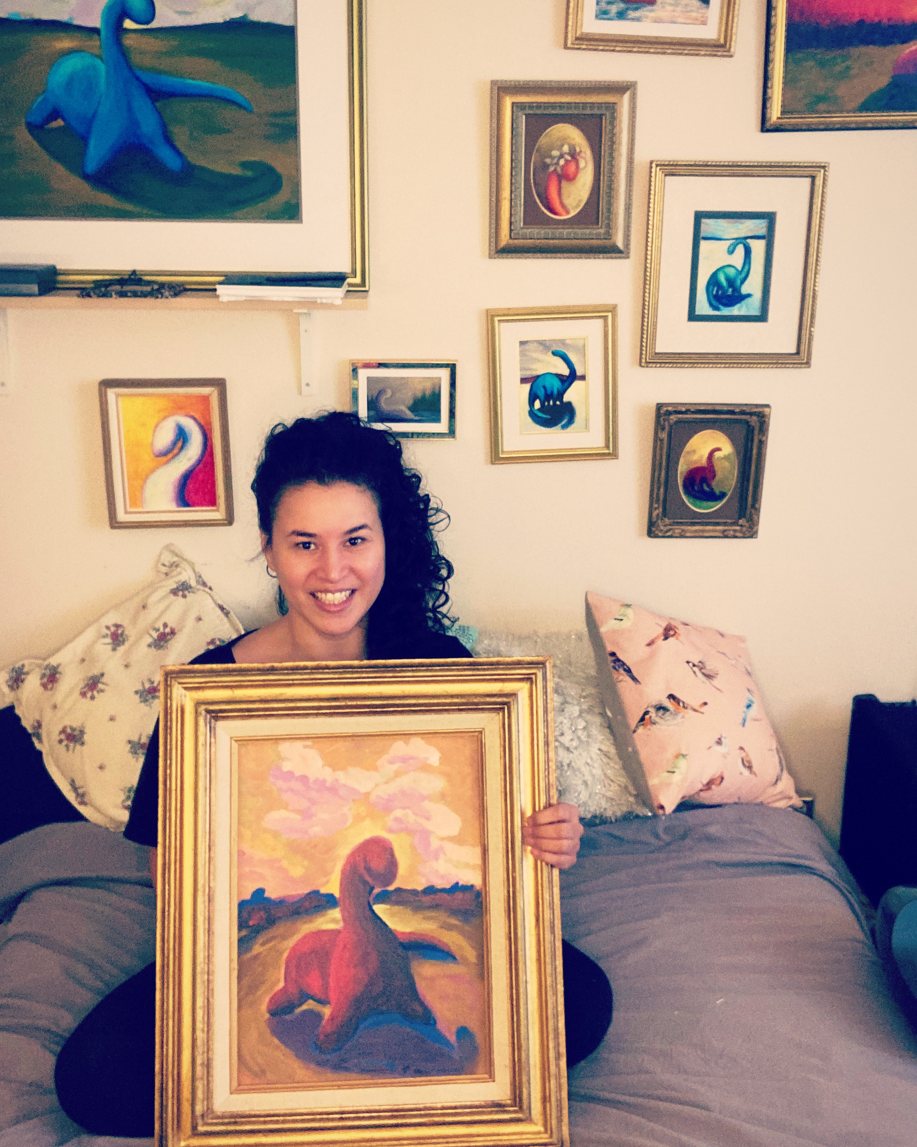 Tiff Hall -very famous artist- holds a framed painting of a dinosaur in front of her humble home studio