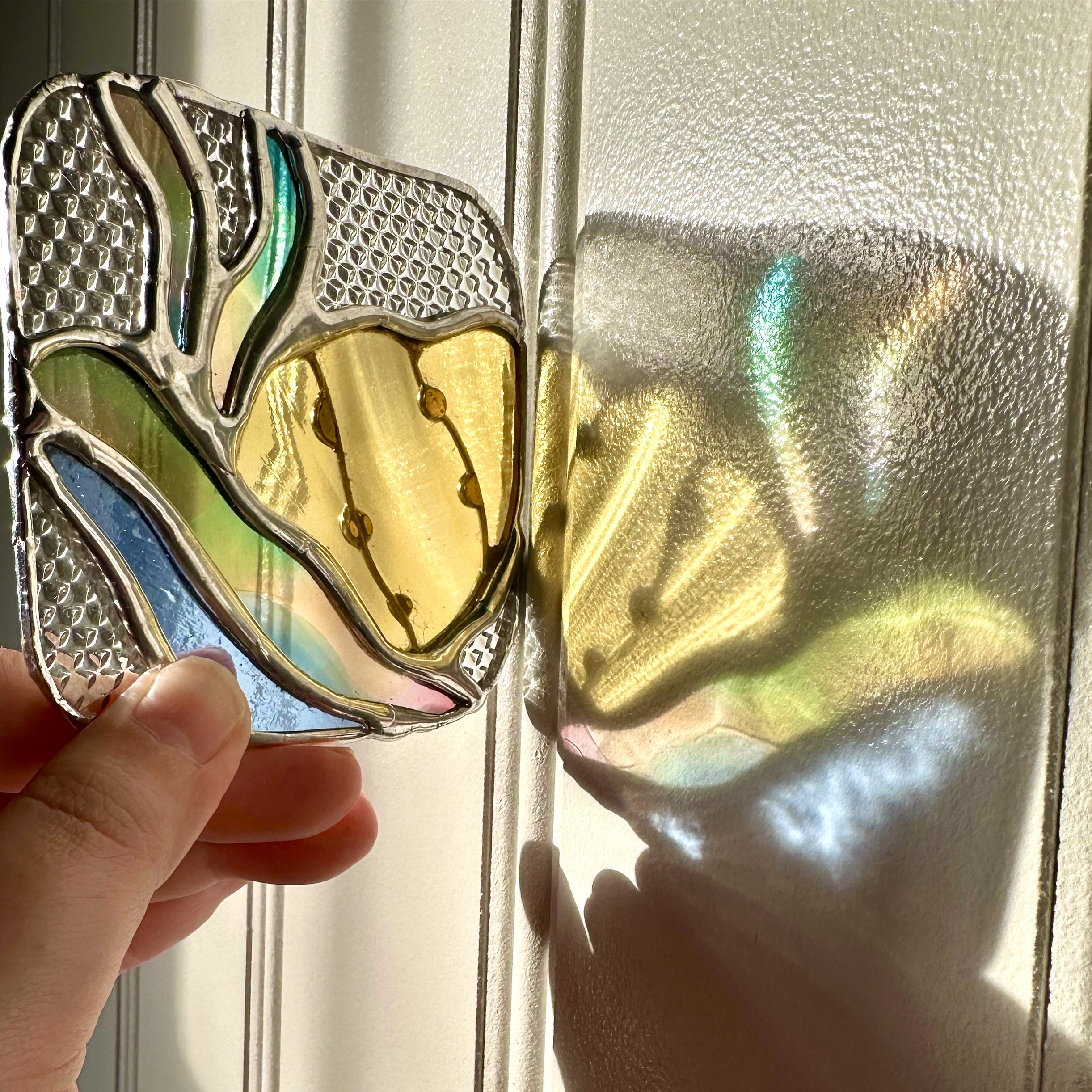 A photo of my completed patch being held in from of a white wall with the sun shining through it to create a colorful refraction on the wall. 