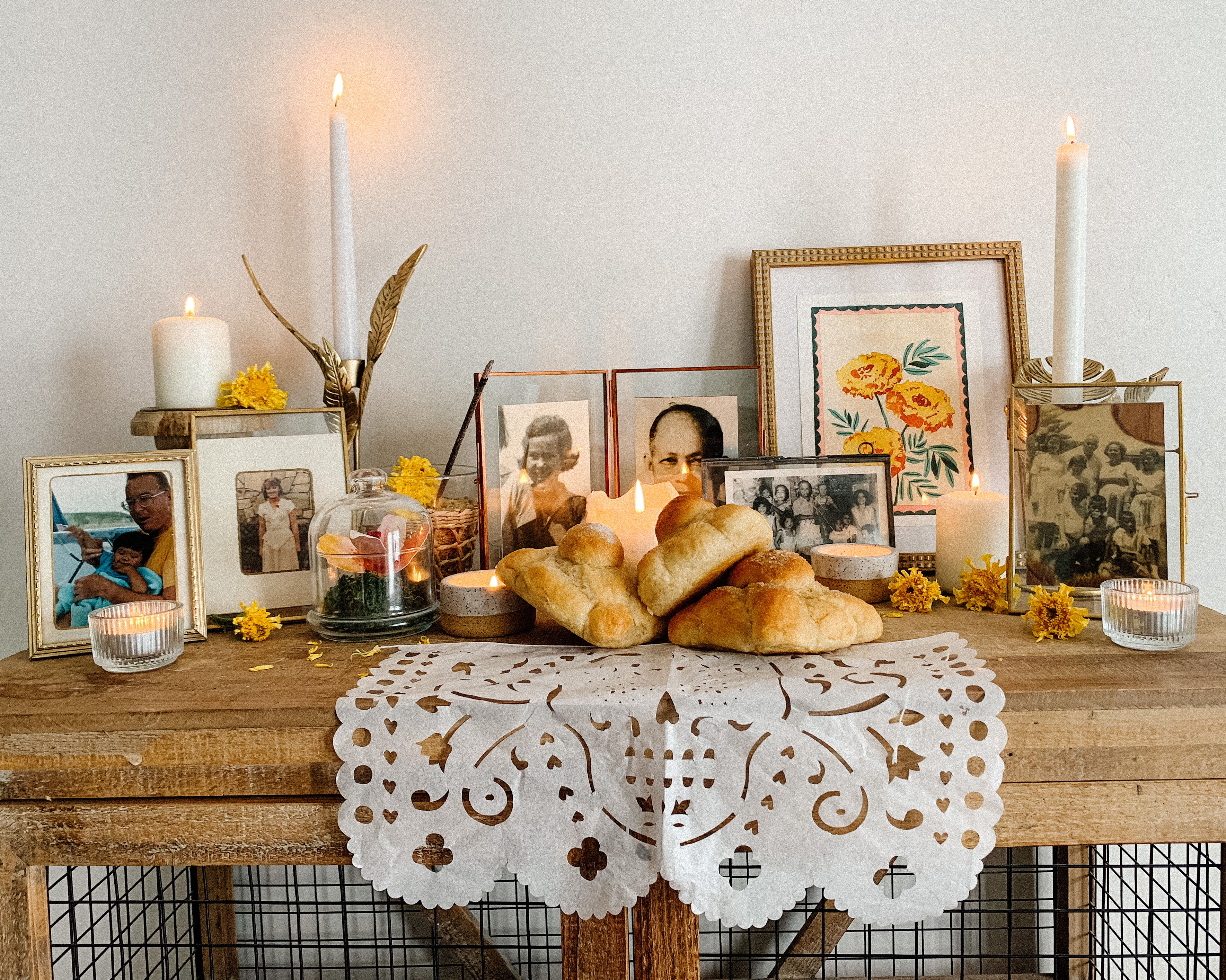 A small ofrenda (or altar) set on top of a bookshelf.  Candles are scattered around, warmly glowing. Sweet rolls, flowers and petals are placed among pictures of family members and friends who have passed.