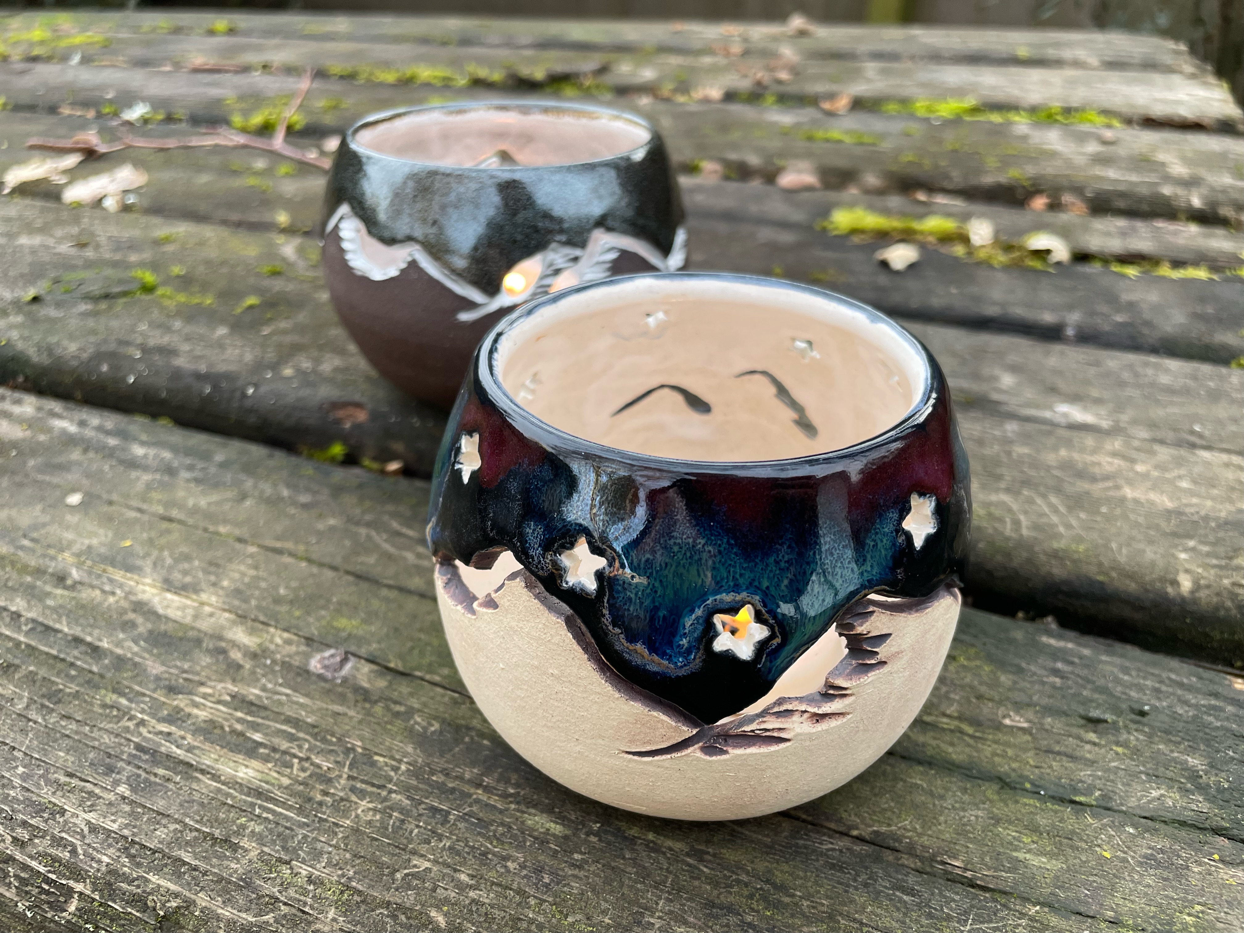 Two round candle holders sit on a table. They are carved with mountains and stars and the sky is glazed to look like the aurora borealis.