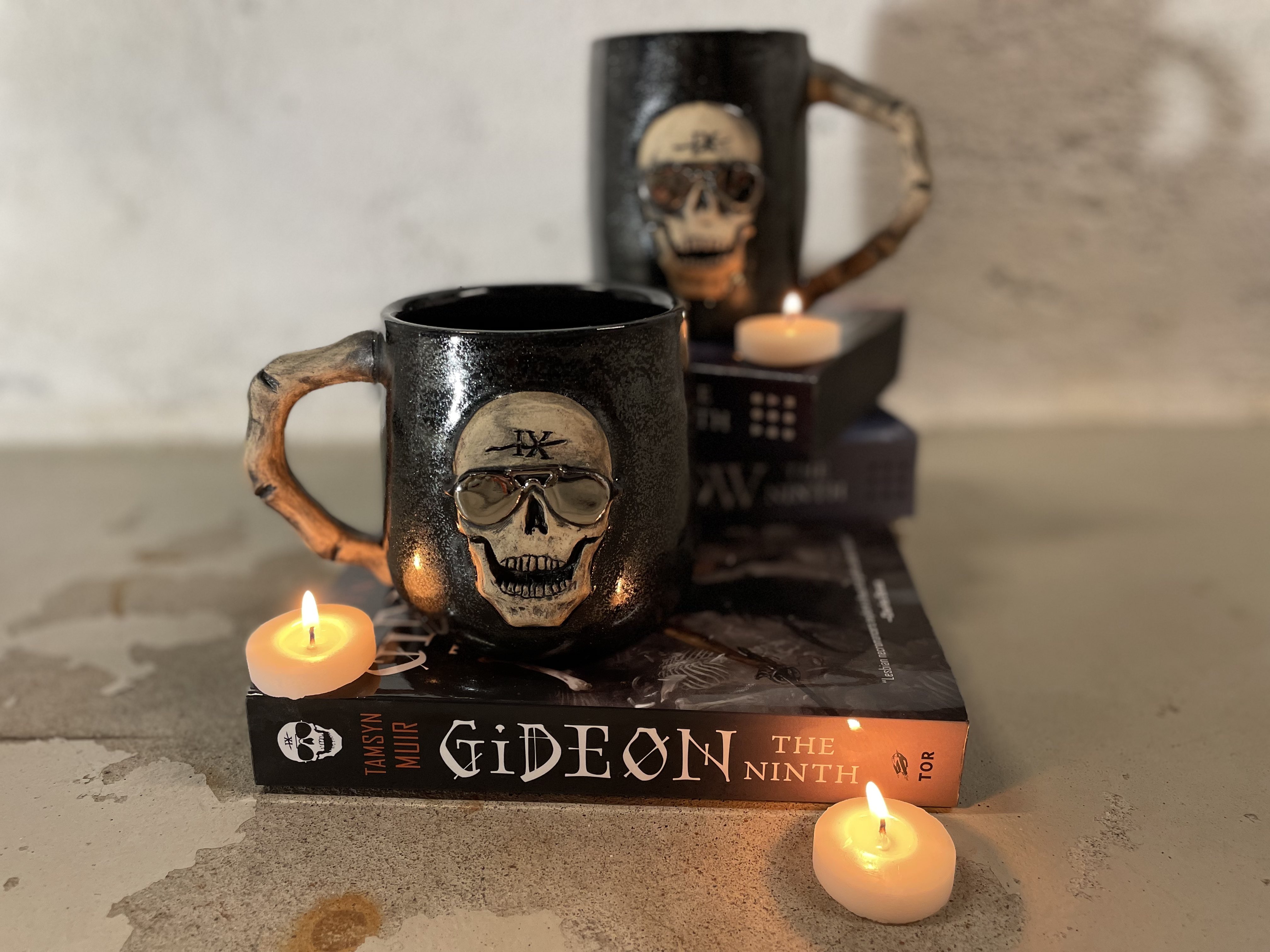 Two black glitter mugs with skulls carved on them and handles that look like finger bones. They are sitting on top of Gideon the Ninth and there are small candles.