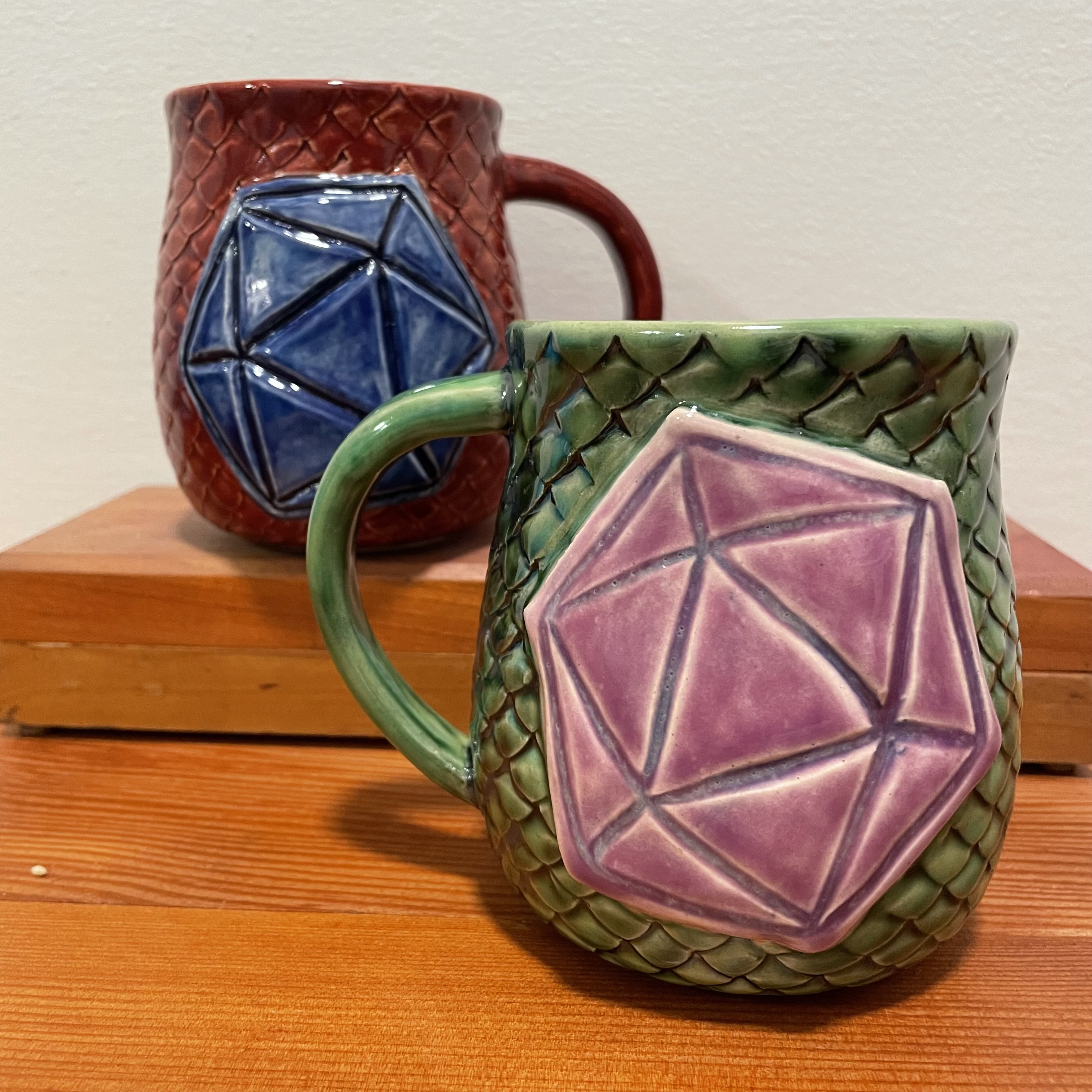 Two mugs with scales carved into them, each has a d20 shape set into them on the front belly of the mug.