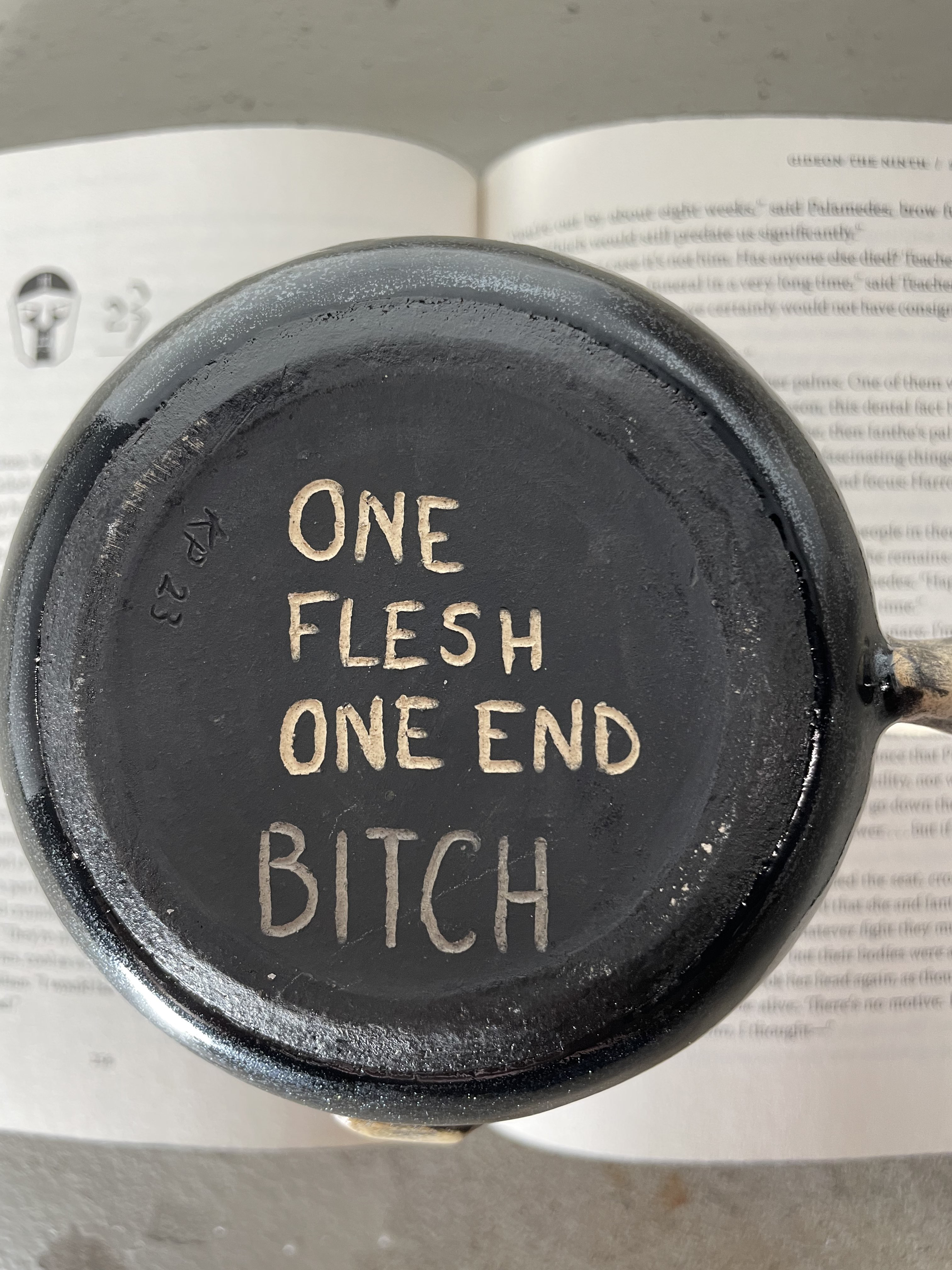 Bottom of a mug, which is carved to read, "One Flesh One End BITCH". Underneath is an open book from the Locked Tomb series.