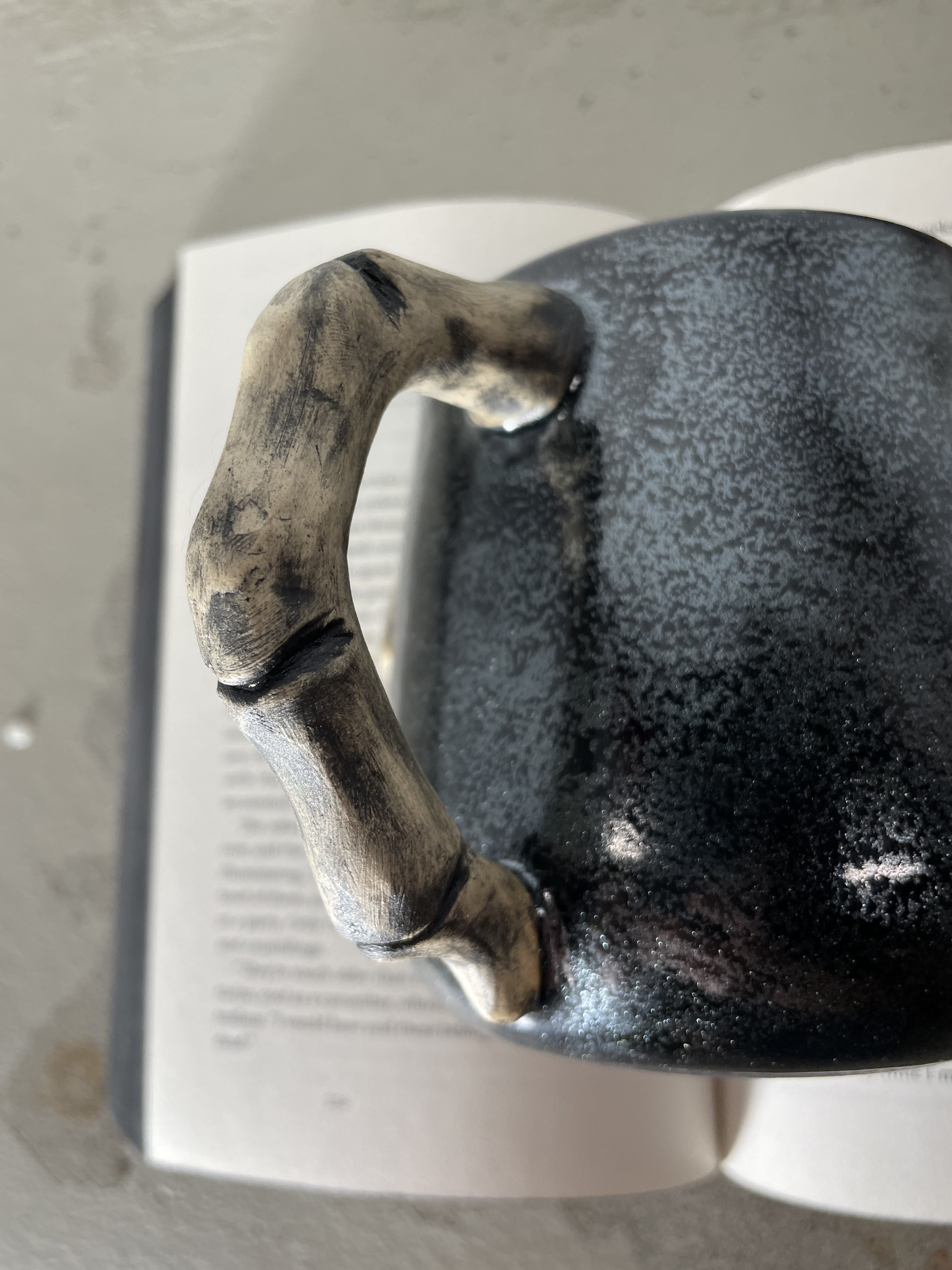 Close up shot of a mug handle shaped like finger bones. In the background is an open book from the Locked Tomb series.