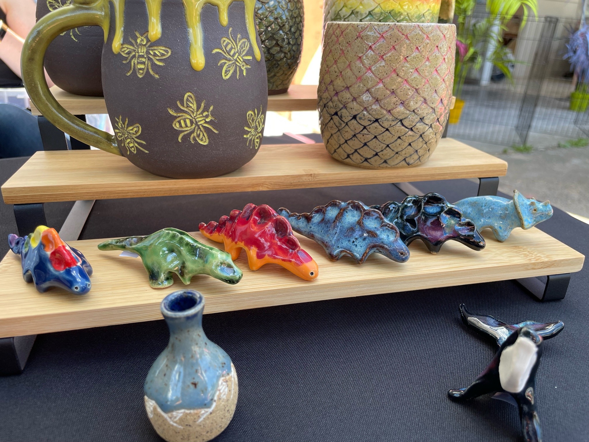 Several rows of ceramics. Back row, brown mug with yellow honey bees and yellow glaze dripping from the rim. Cup with carved scales, with background of pink, purple and blue. Second row, assorted small dinosaurs. Front row, small blue and white vase and a small orca.