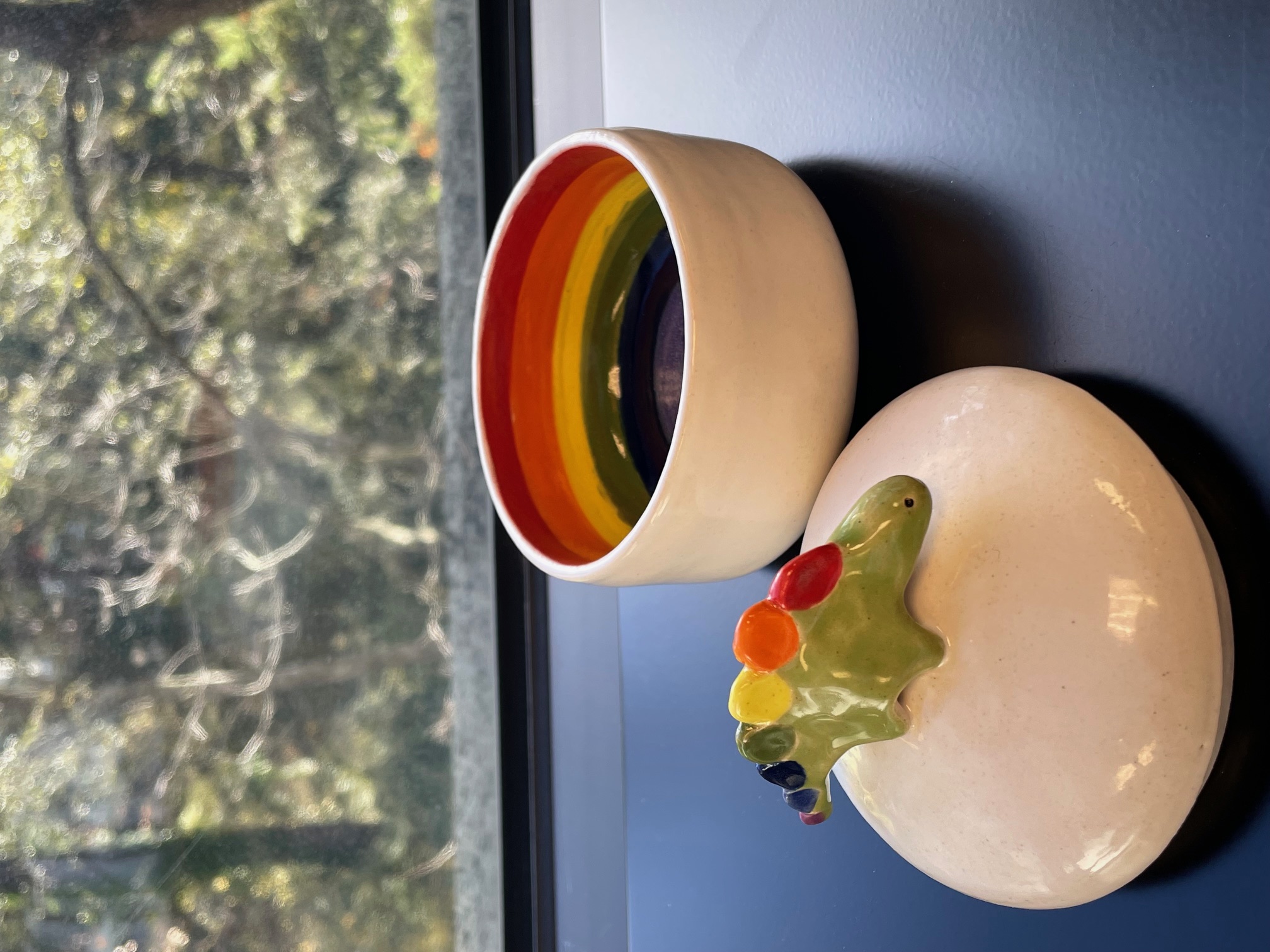 White trinket bowl with lid off to the side. The lid has a stegosaurus with painted rainbow spines. The inside of the bowl is painted in a rainbow.