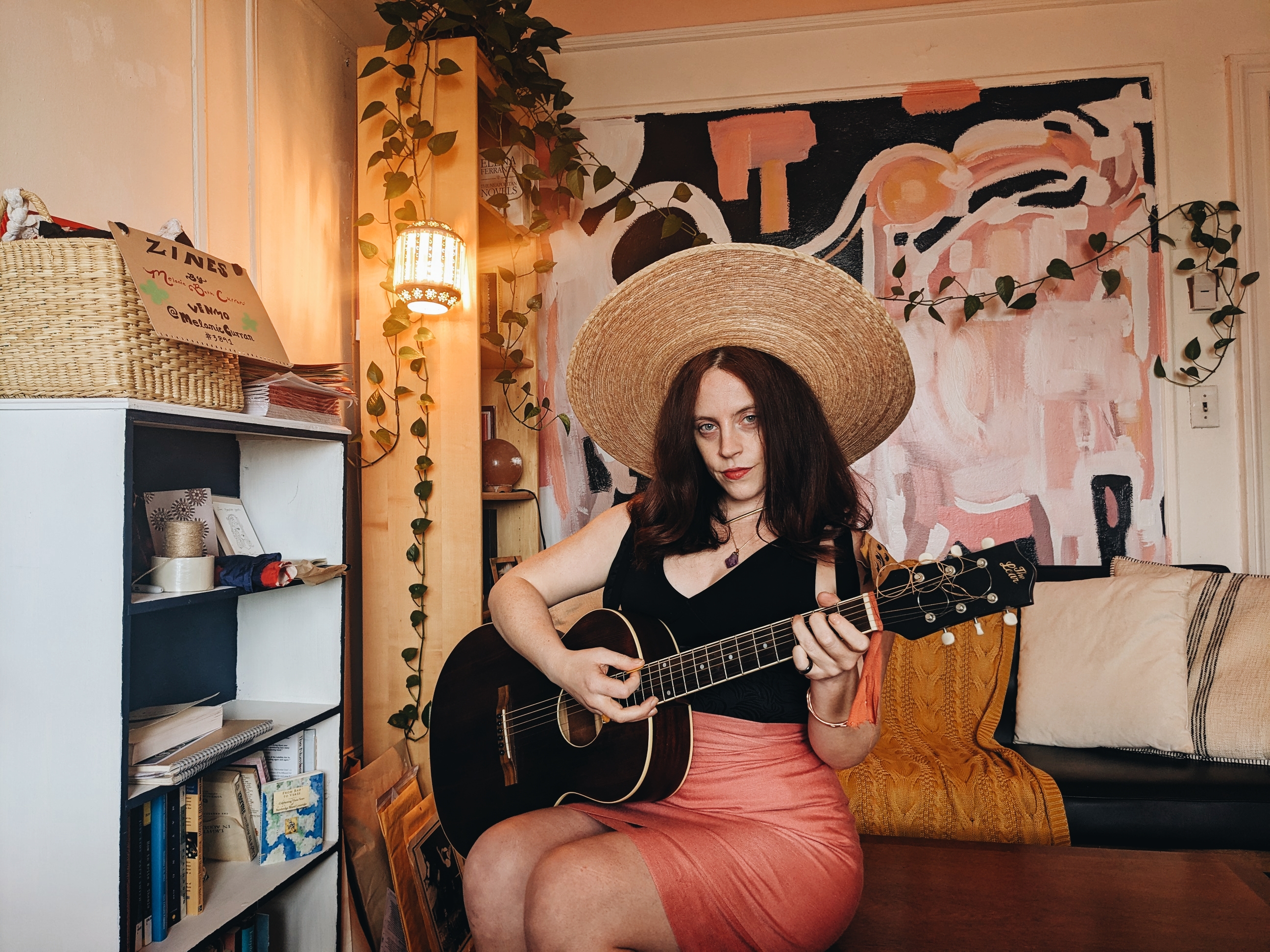 Melanie Beth Curran wears a large straw hat and plays a parlor guitar in her scriptorium, she has auburn hair and green eyes