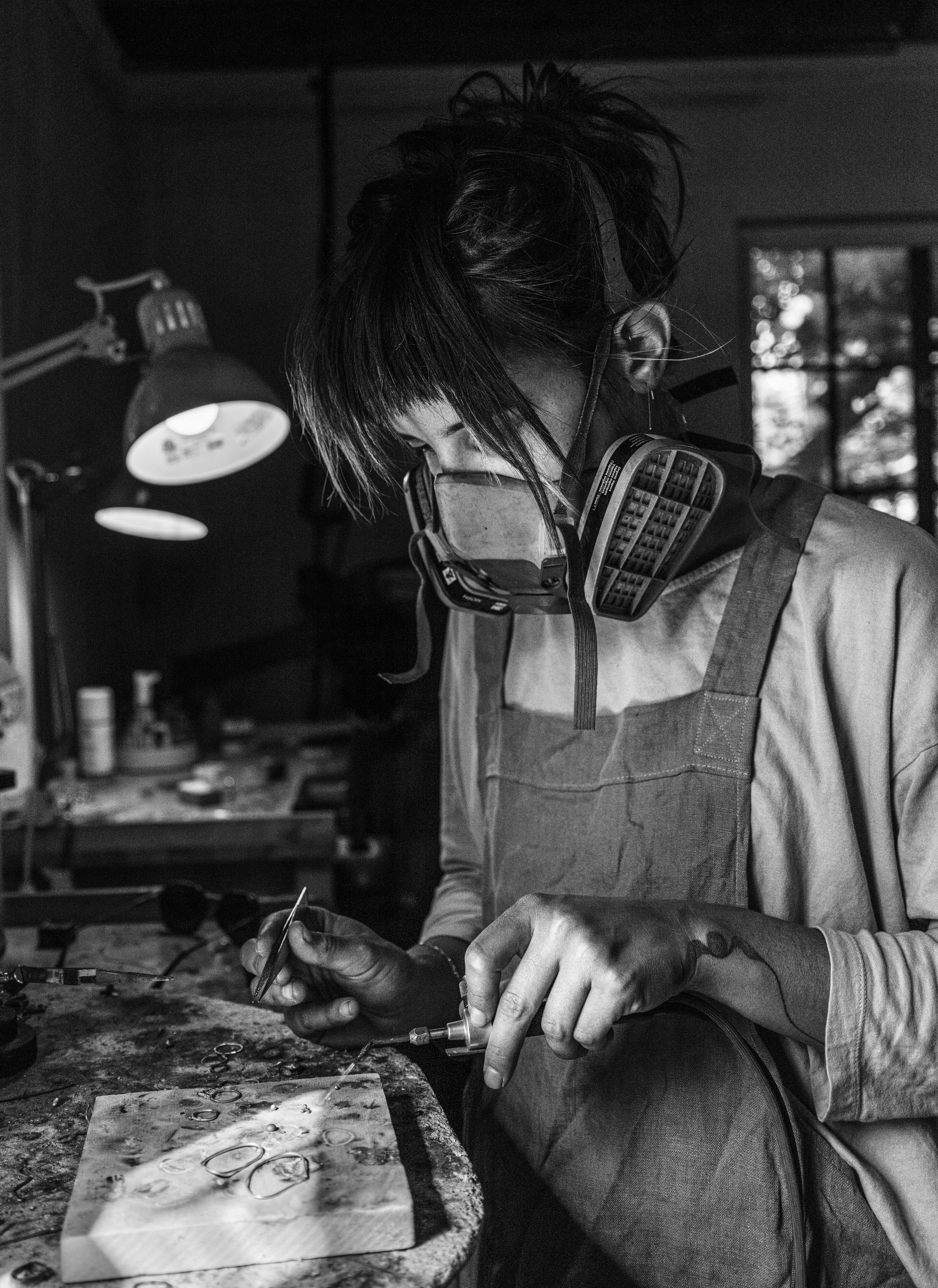 Me, Devon Gray, in black and white, a respirator, and torch in my metalsmithing studio, in Louisville Kentucky. 