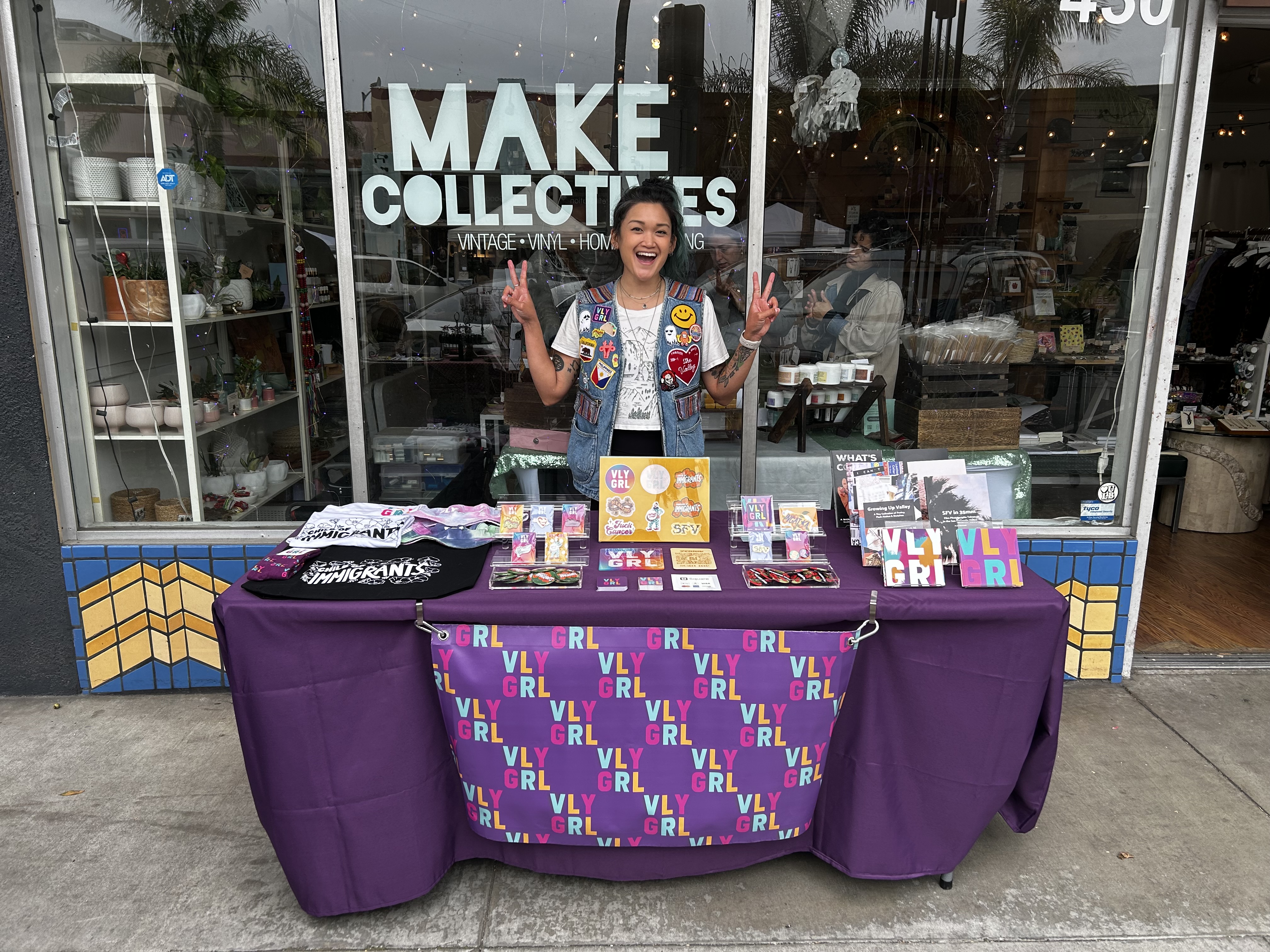 Alisa, founder of VLY GRL, stands behind a vending table at a storefront. Various VLY GRL merchandise and zines sit on the table while Alisa, wearing a jean vest with patches on it, is smiling while making peace signs with her hands.