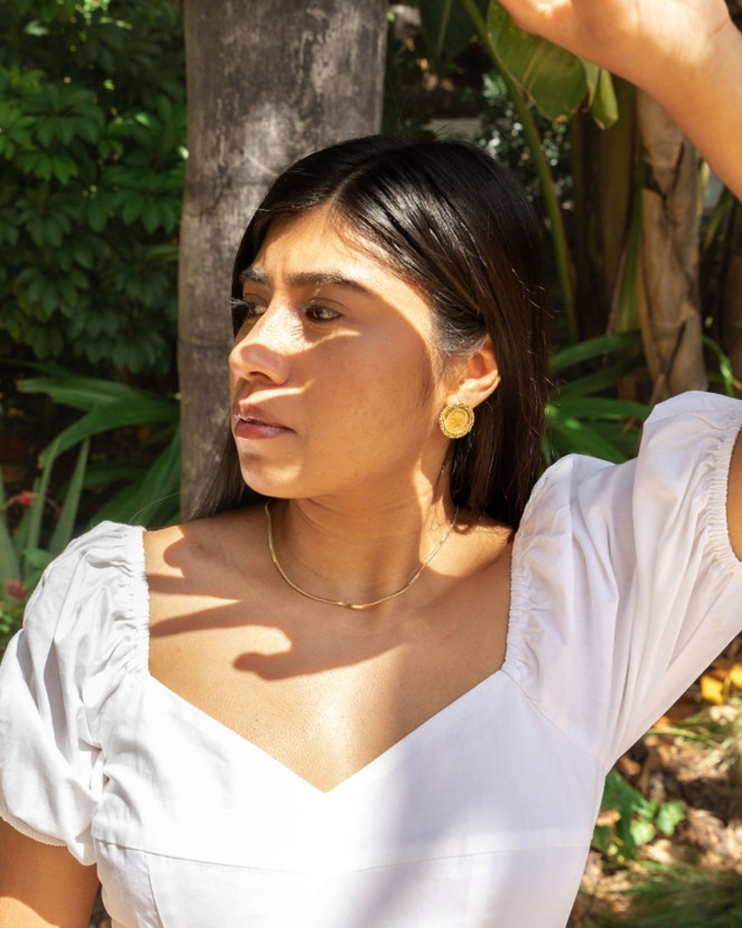 Diana Pimentel, the founder of Piel Culta, stands outdoors in a garden with lush greenery in the background. She is wearing a white dress with puffed sleeves, gold earrings, and a delicate gold necklace. She has long dark hair and is partially shaded, with sunlight creating dappled patterns on her face and dress. Her expression is thoughtful as she looks off into the distance, with one hand raised slightly to shield her eyes from the sun.