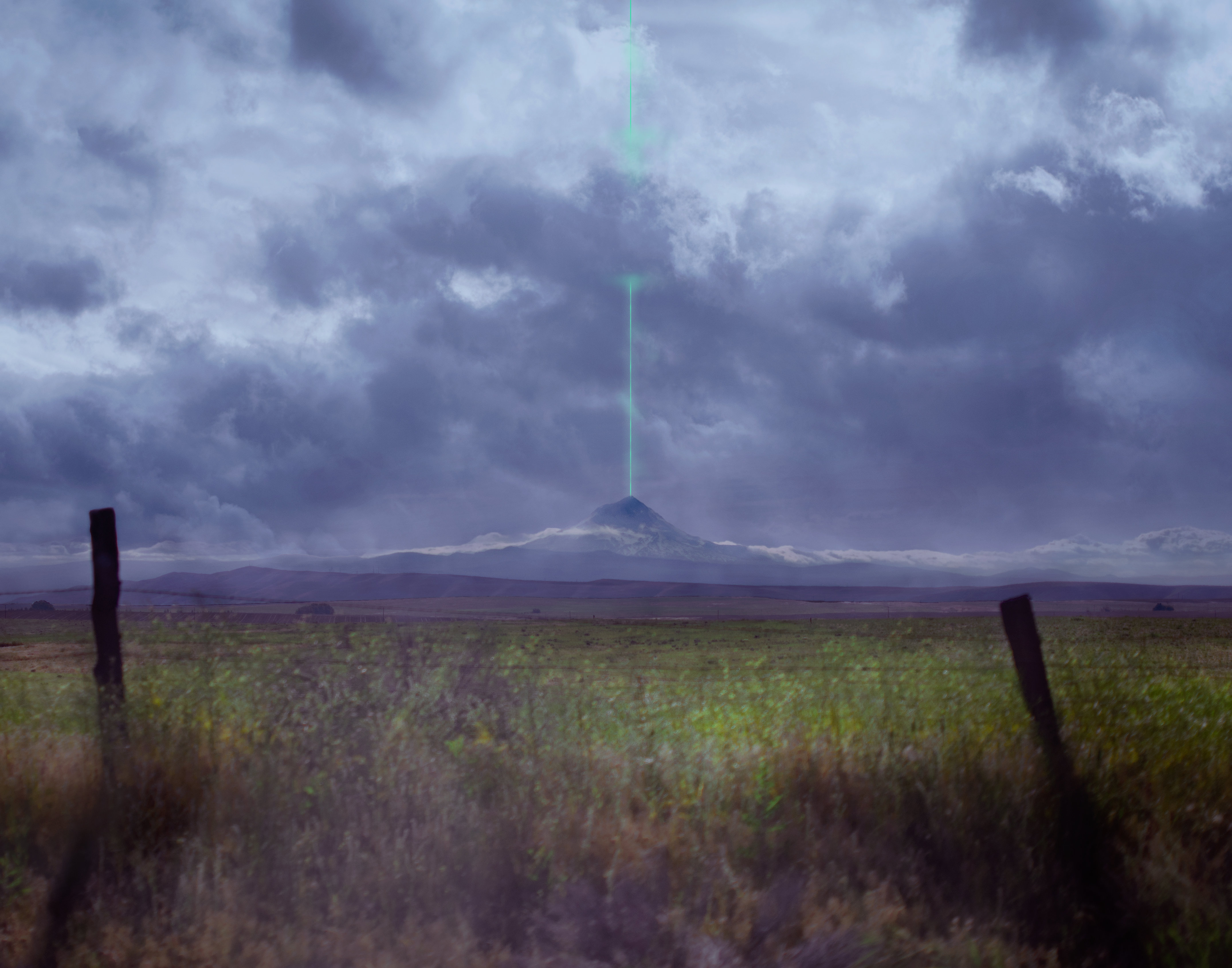 A color photograph of Mt. Hood in the distance. It stands under a purple sky filled with clouds. in the foreground, a murky green field fills the distance between the viewer and the mountain, and two black fence poles stand on either side of the frame, on the edge of the field closest to the viewer. From the top of Mt Hood a bright teal laser or light beam emits straight up into the clouds.