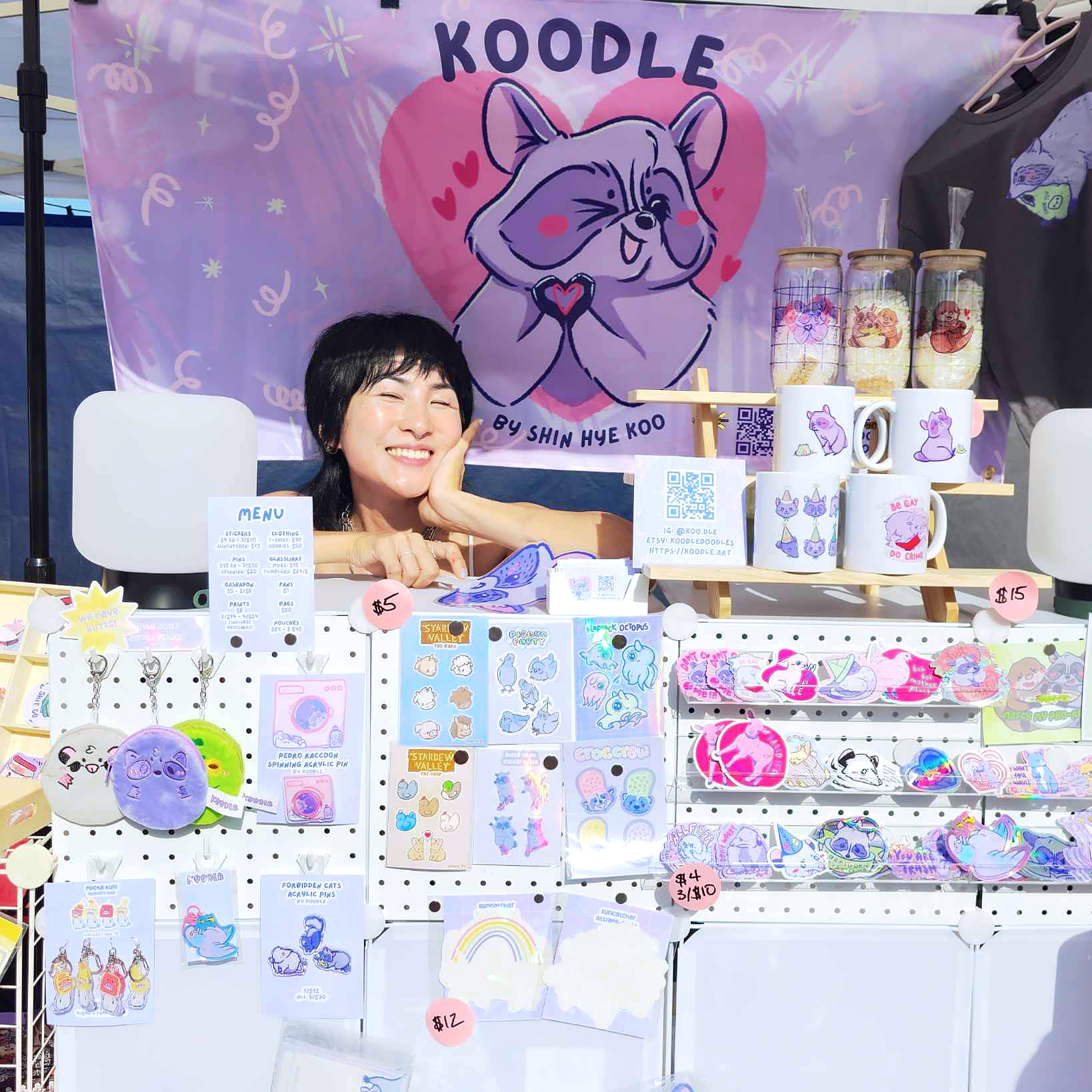 A photo of Shin Hye Koo, the artist behind Koodle, standing behind her display. Behind her is a banner featuring her original character, Kuni, who is signing a heart with their hands.