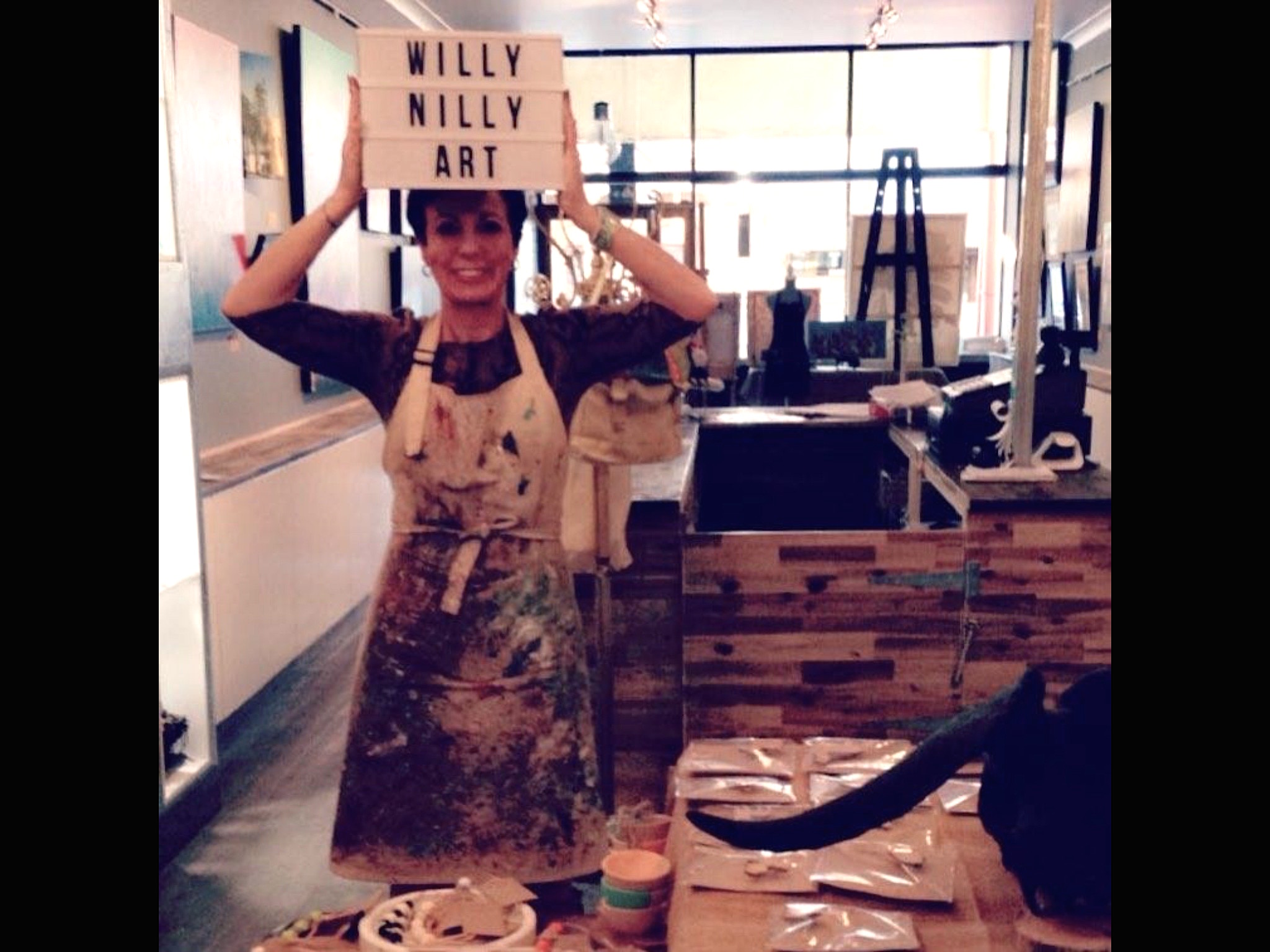 The Artist Amanda Johnson holds up a sign that says Willy Nilly Art. The background is the retail space of her store showing original artworks, jewellery and an apron