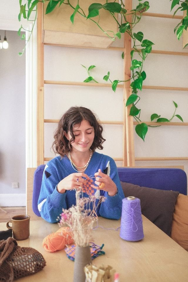 Woman crocheting in front of a wood backdrop with hanging plants. She is smiling and has brown curly hair.