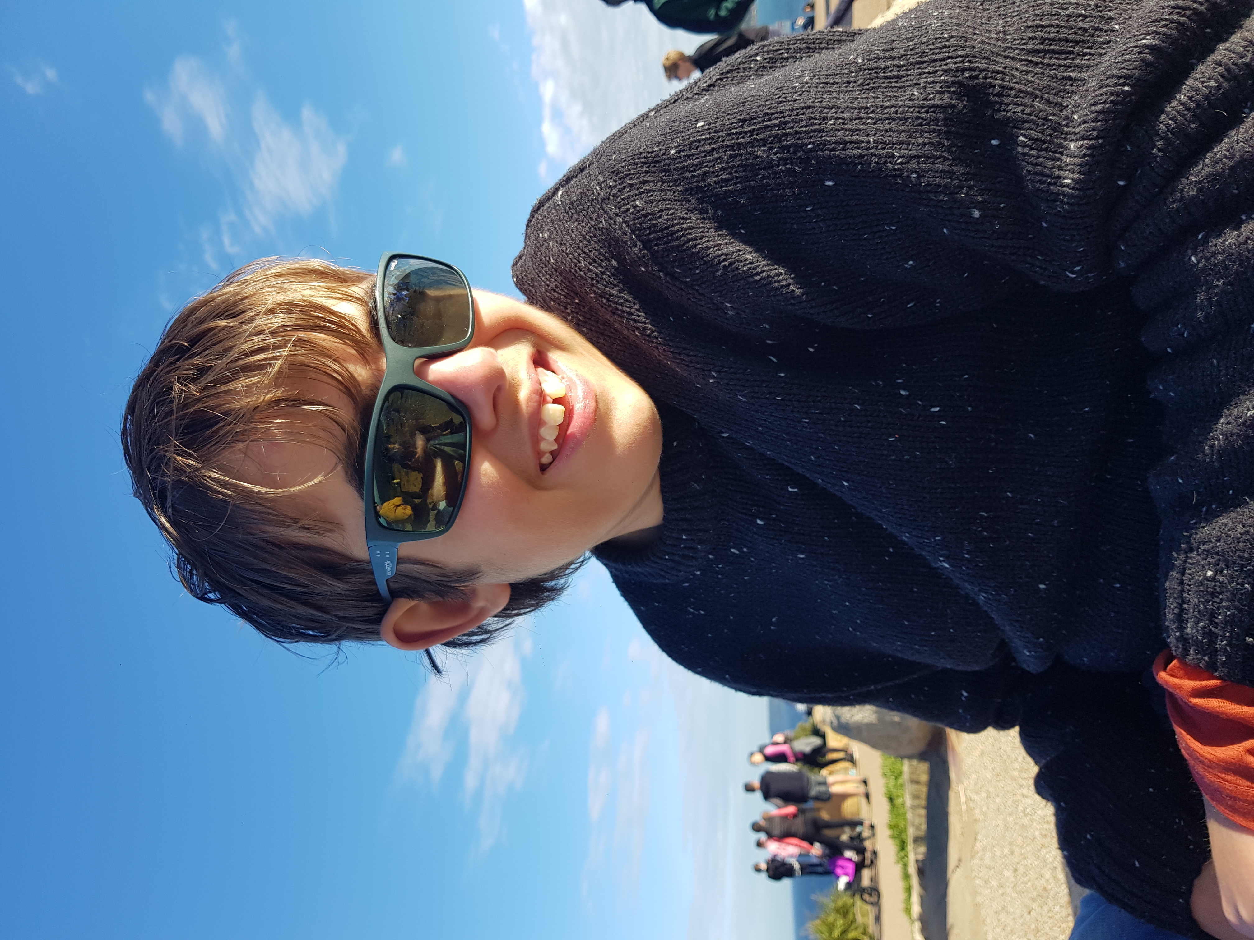 A photo of Luca wearing a dark sweater and sunglasses, sitting in the sun. The sky is bright blue behind him, and he is smiling at the camera.