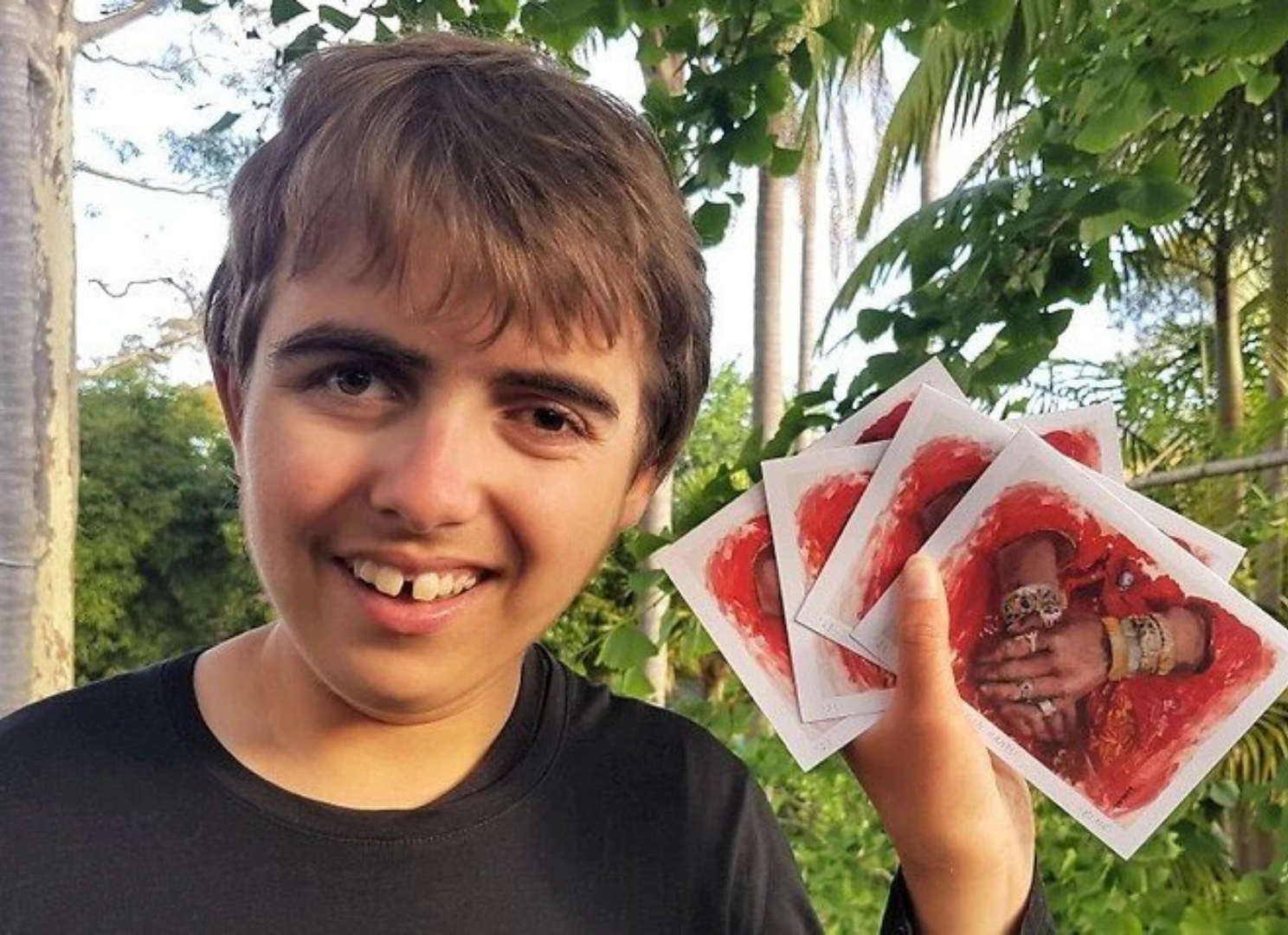 A photo of Luca holding 4 red Afghanistan fundraising cards in his left hand. He is wearing a dark shirt and is smiling at the camera, and is in front of some leafy trees.