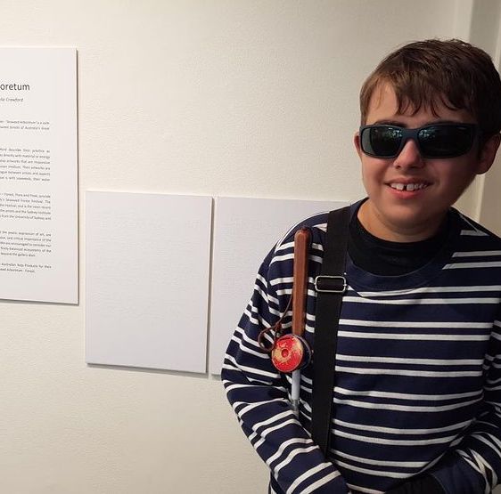 A photo of Luca in front of 2 brailled panels, next to the artwork description. Luca is smiling at the camera, and is wearing a striped shirt and holding a white cane. 