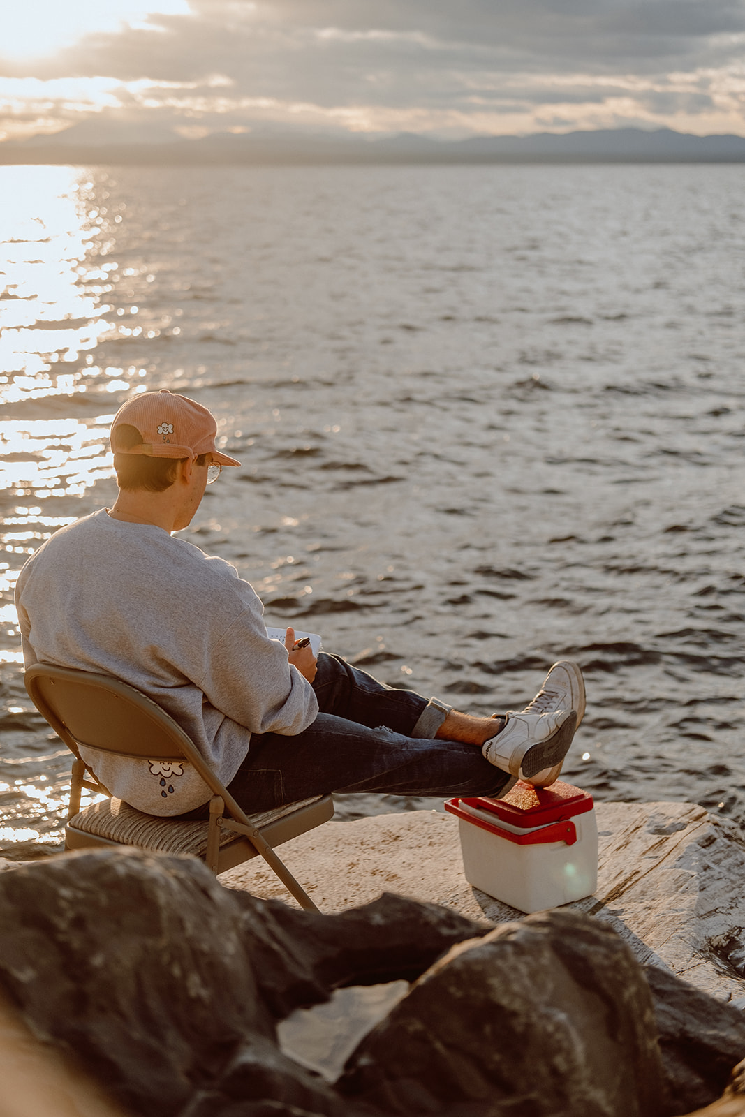 Man sitting in front of lake drawing during sunset.