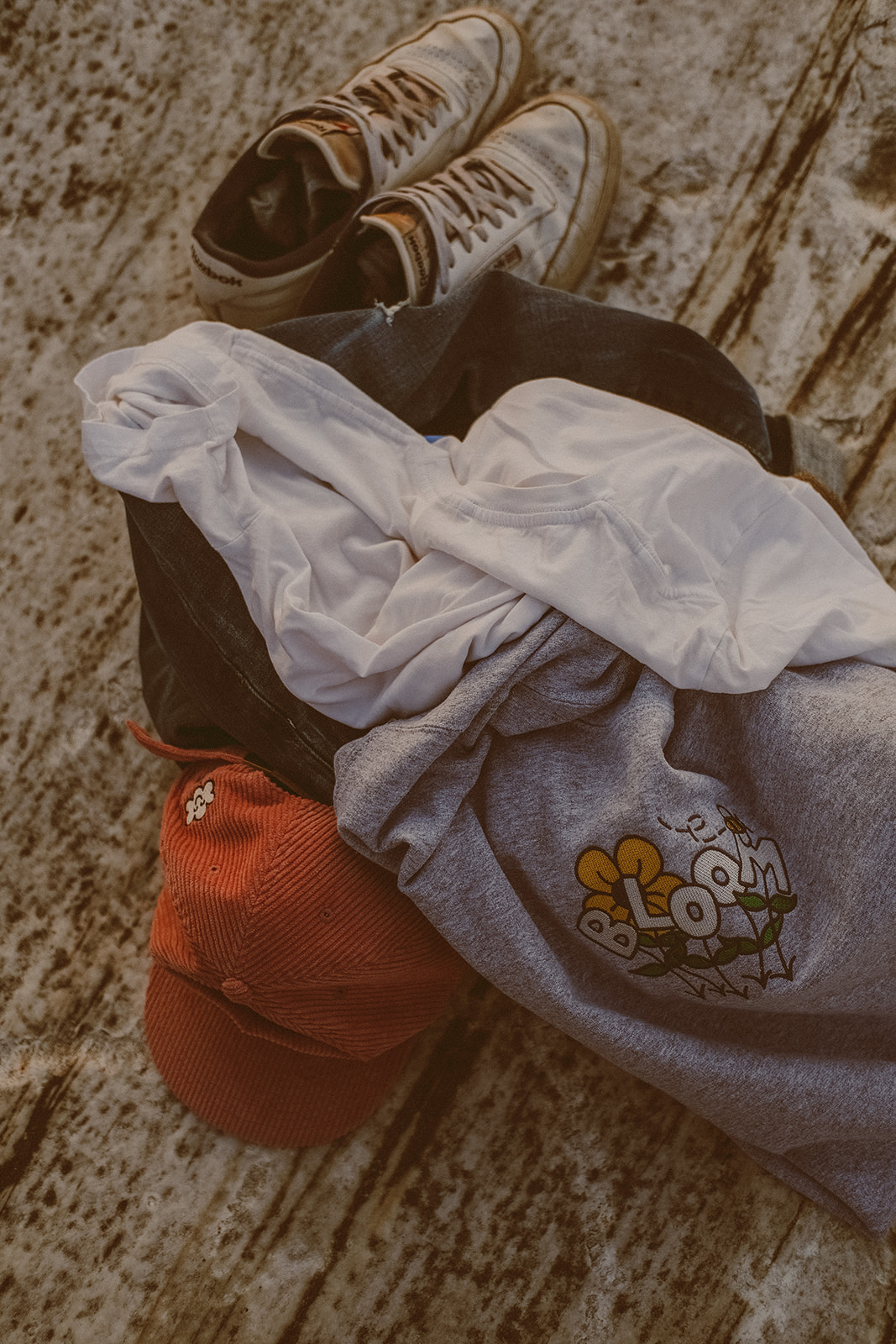 Pile of clothing with white sneakers, blue jeans, white t-shirt, grey crewneck and pink corduroy hat sitting on top of a rock.