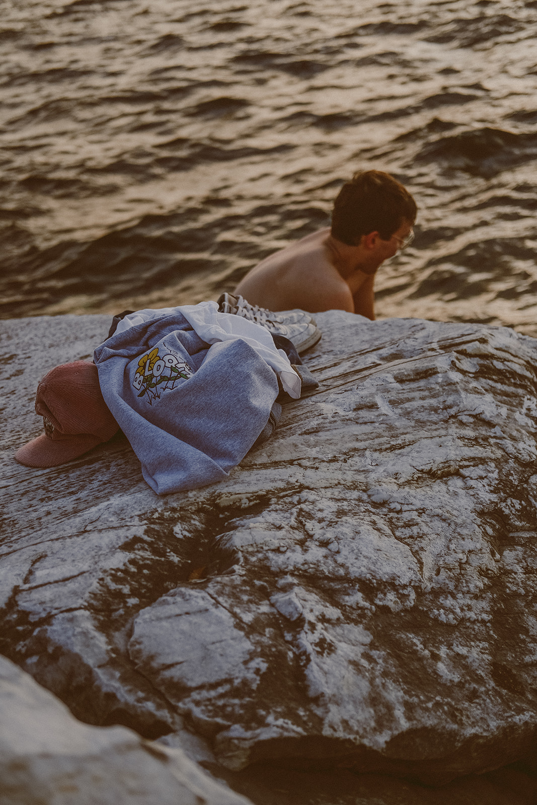 Pile of clothes sitting on a rock in front of the water with a shirtless man visible behind the rock in the water.
