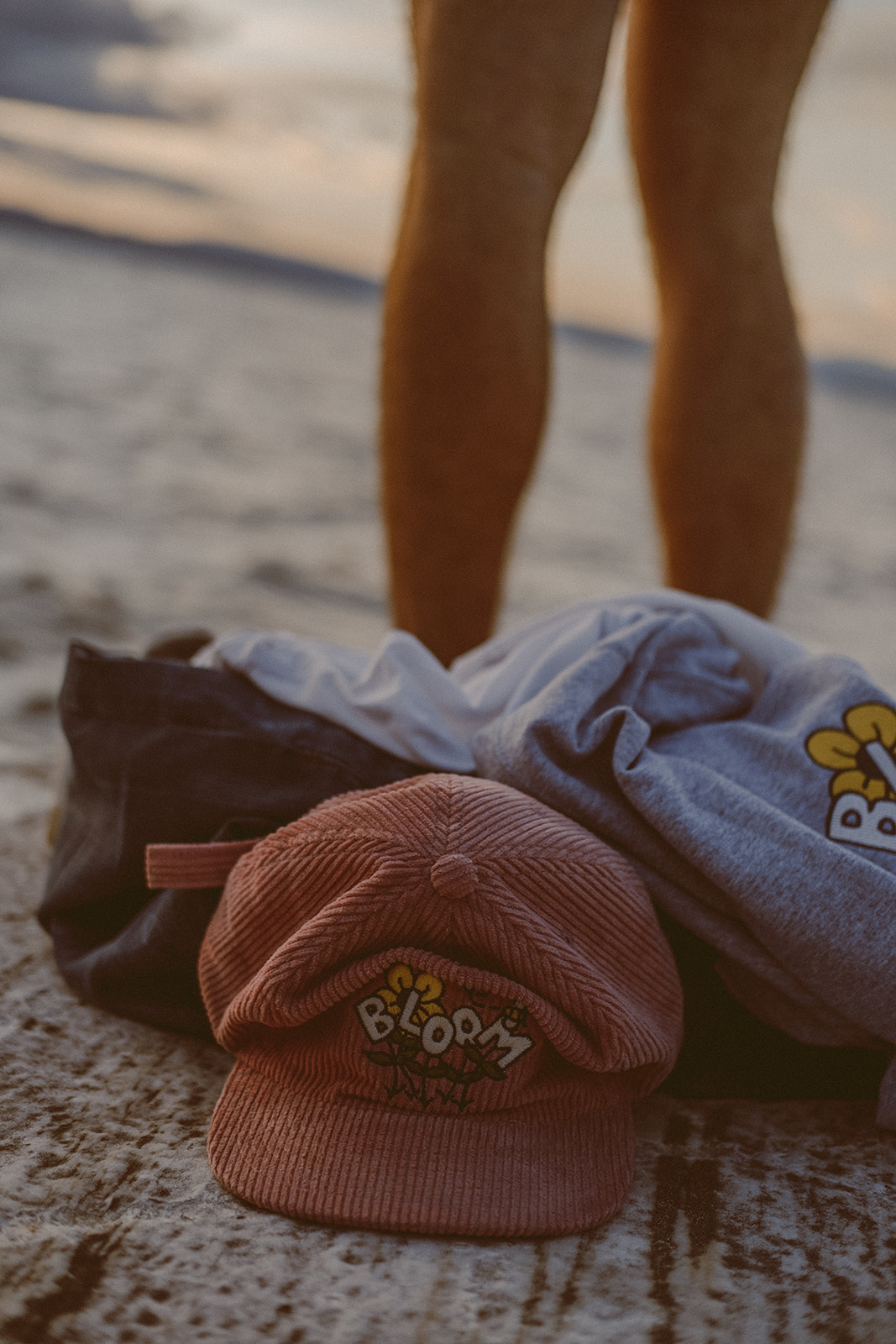 Pink corduroy hat with "BLOOM" logo sitting in front of a pile of clothes with a mans bare legs in the background.
