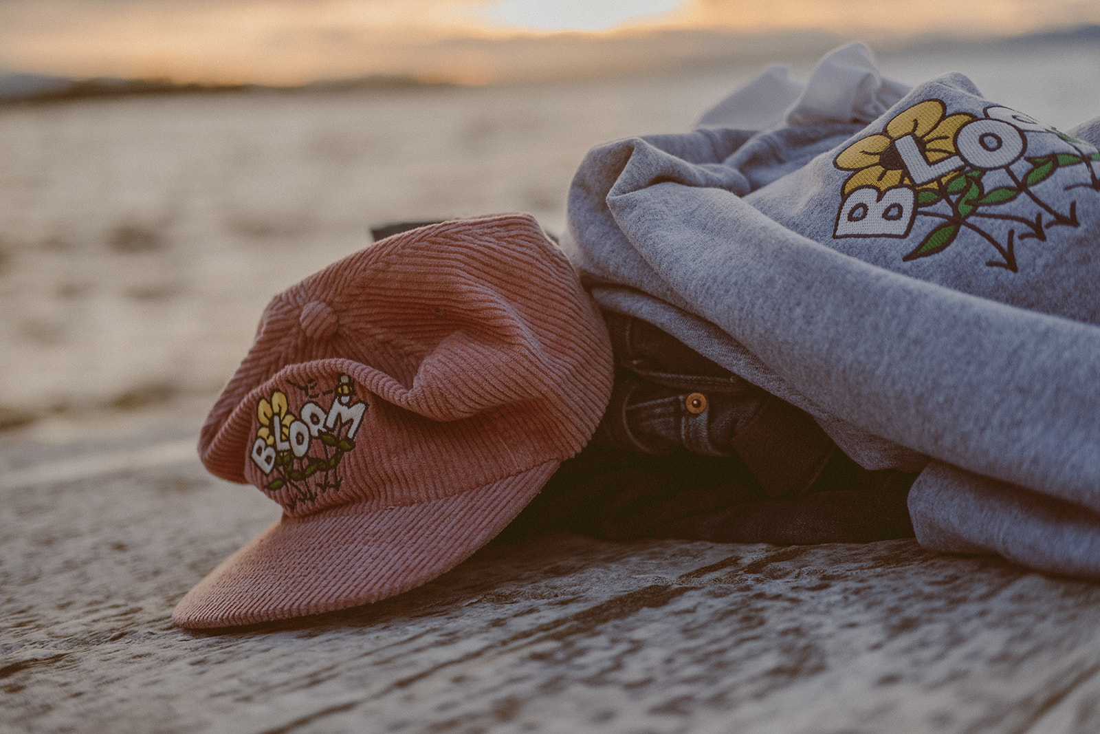 Pile of clothes with pink corduroy hat and grey crewneck with "BLOOM" logo sitting on top in front of a sunset.