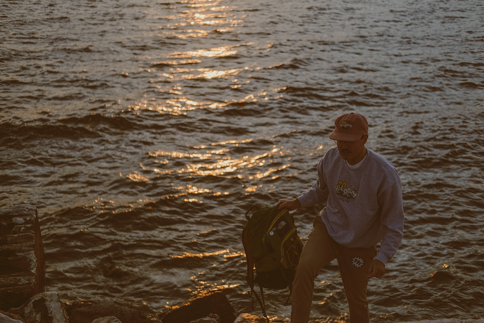 Man in front of the sunset reflecting off of a lake carrying a backpack in one hand wearing a pink hat, grey crewneck and tan pants.