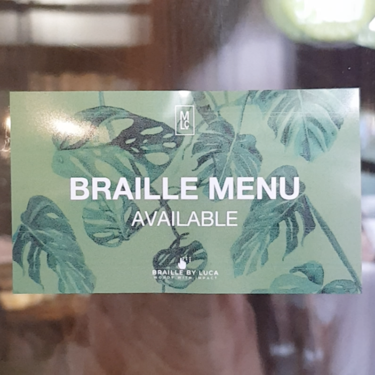An image of a business card that says "Braille Menu Available" with the Market Lane Cafe logo andthe Braille by Luca logo. The text and logos are white, and the background are green leaves. 