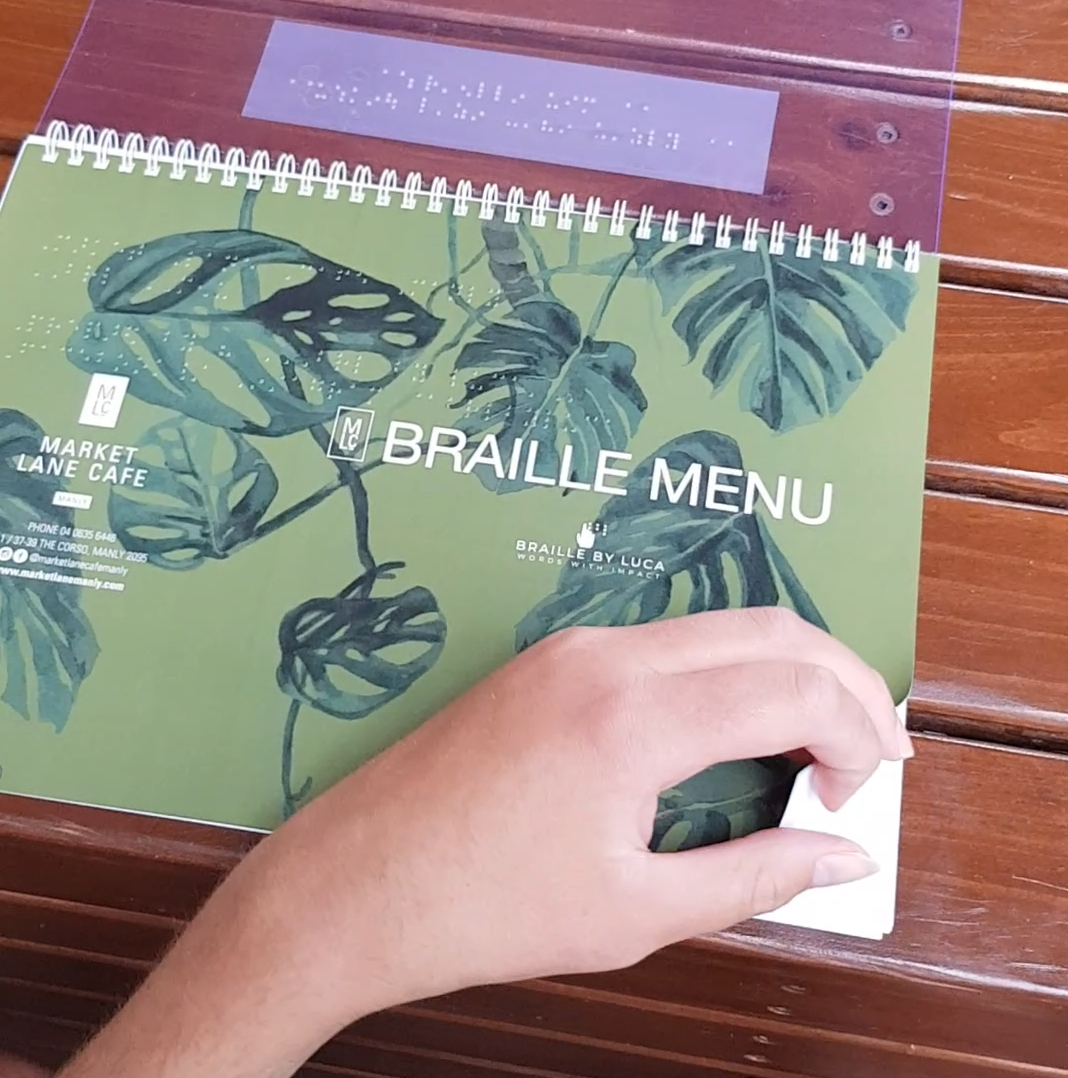 An image of the front page of the braille Market Lane cafe menu. It says "Braille Menu" and includes both the Braille by Luca and Market Lane Cafe logos. It is white text on a green, leafy background. Lucas hand is turning the page. 