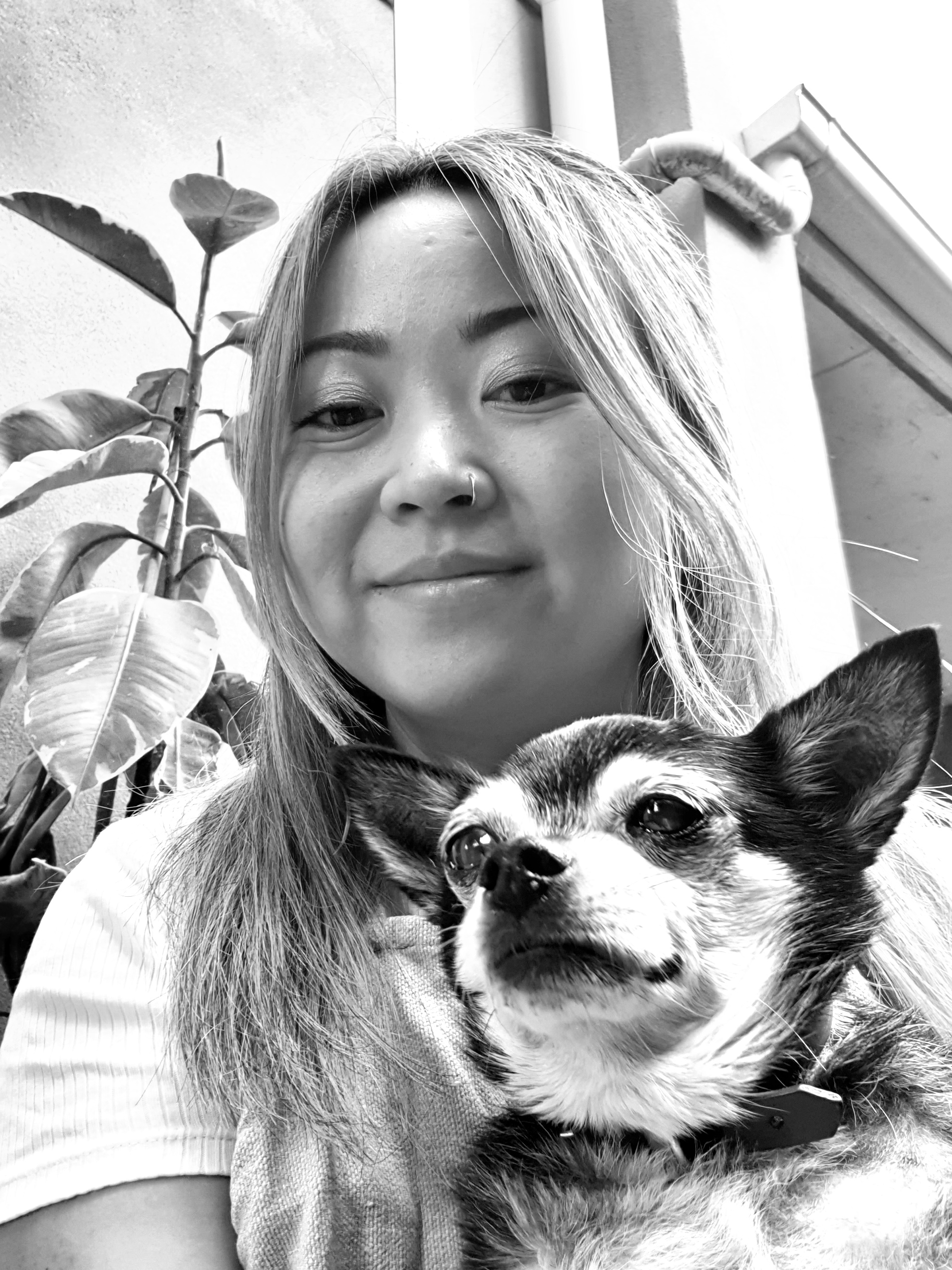 B;ack and white image of a woman with long hair half pulled up smiling holding a chihuahua with a plant and building in the background.