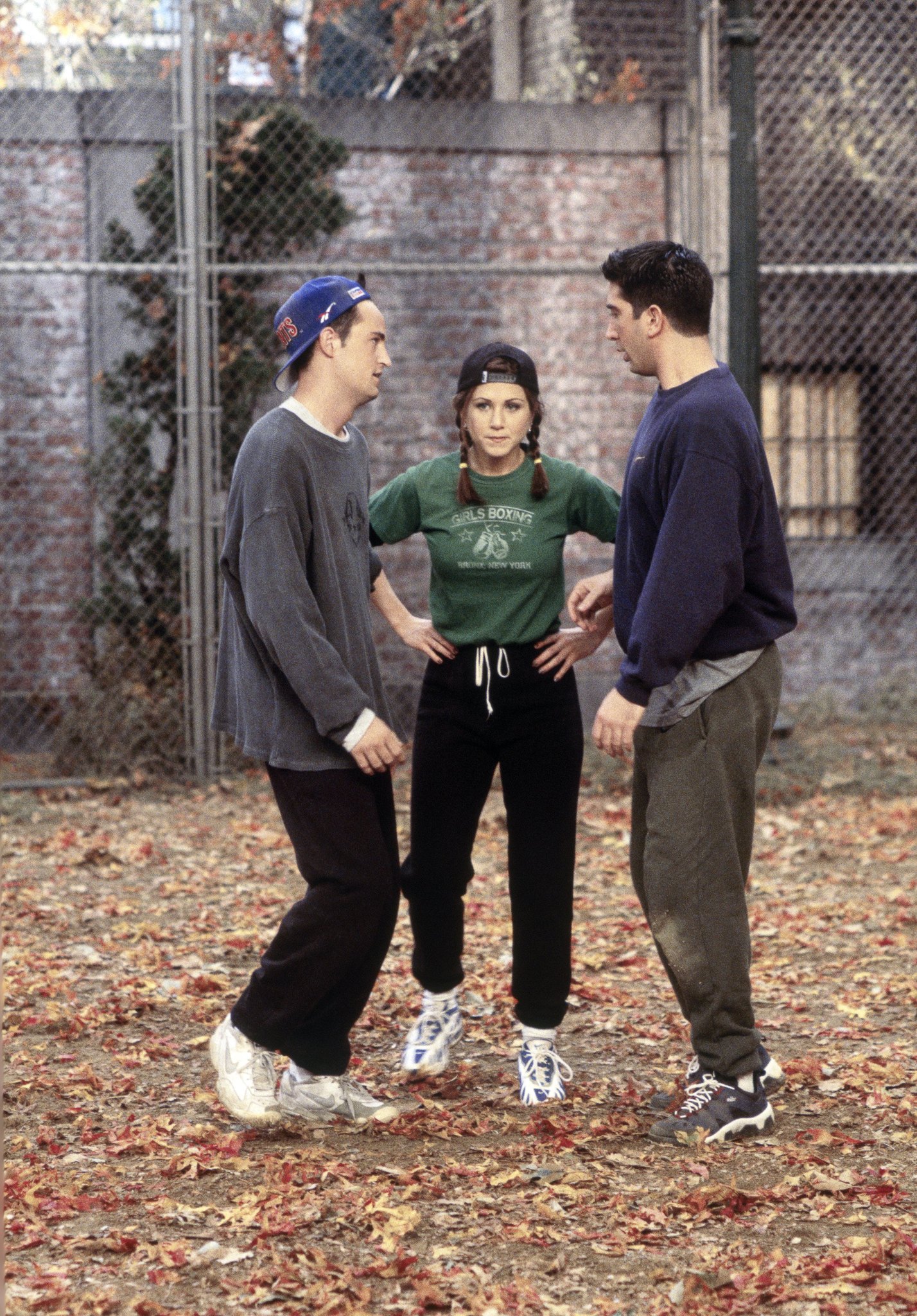 Chandler, Rachel and Ross from FRIENDS stood together in a line. Rachel has hands on her hips wearing a cap backwards with plaits in, a long sleeved black t-shirt with a green graphic t-shirt over the top, black jog pants and white trainers. Background is a leafy autumn park scene.