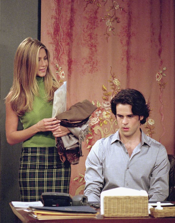 Rachel Green in a green sleeveless rollback jumper with a checkered skirt holding files whilst looking at her male assistant