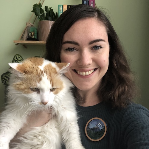 A woman with wavy brown hair holds up a fluffy white and ginger cat. She's smiling and the cat looks grumpy. She's wearing a dark green top and a brooch depicting a mountain scene.