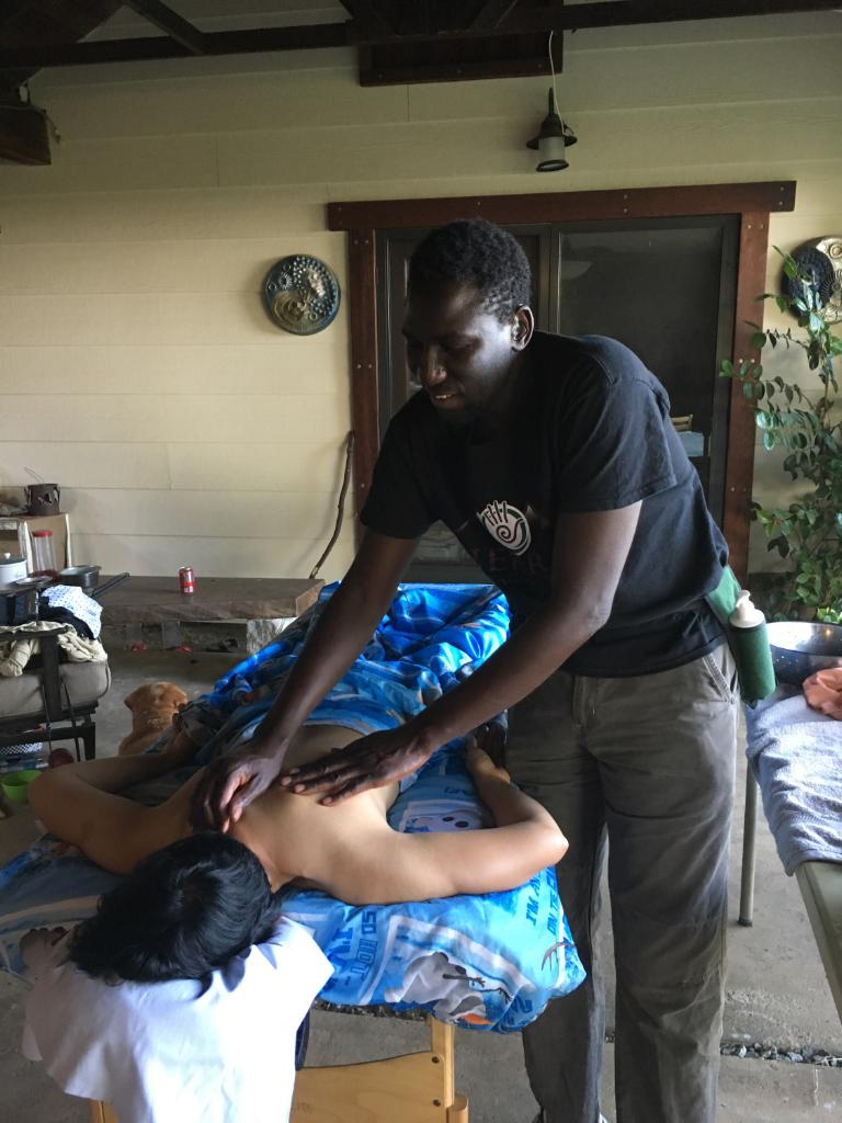 C.M.T. Yegue B providing Massage Therapy on someone's shoulders; person is lying on Table faced down, under a light blanket. 