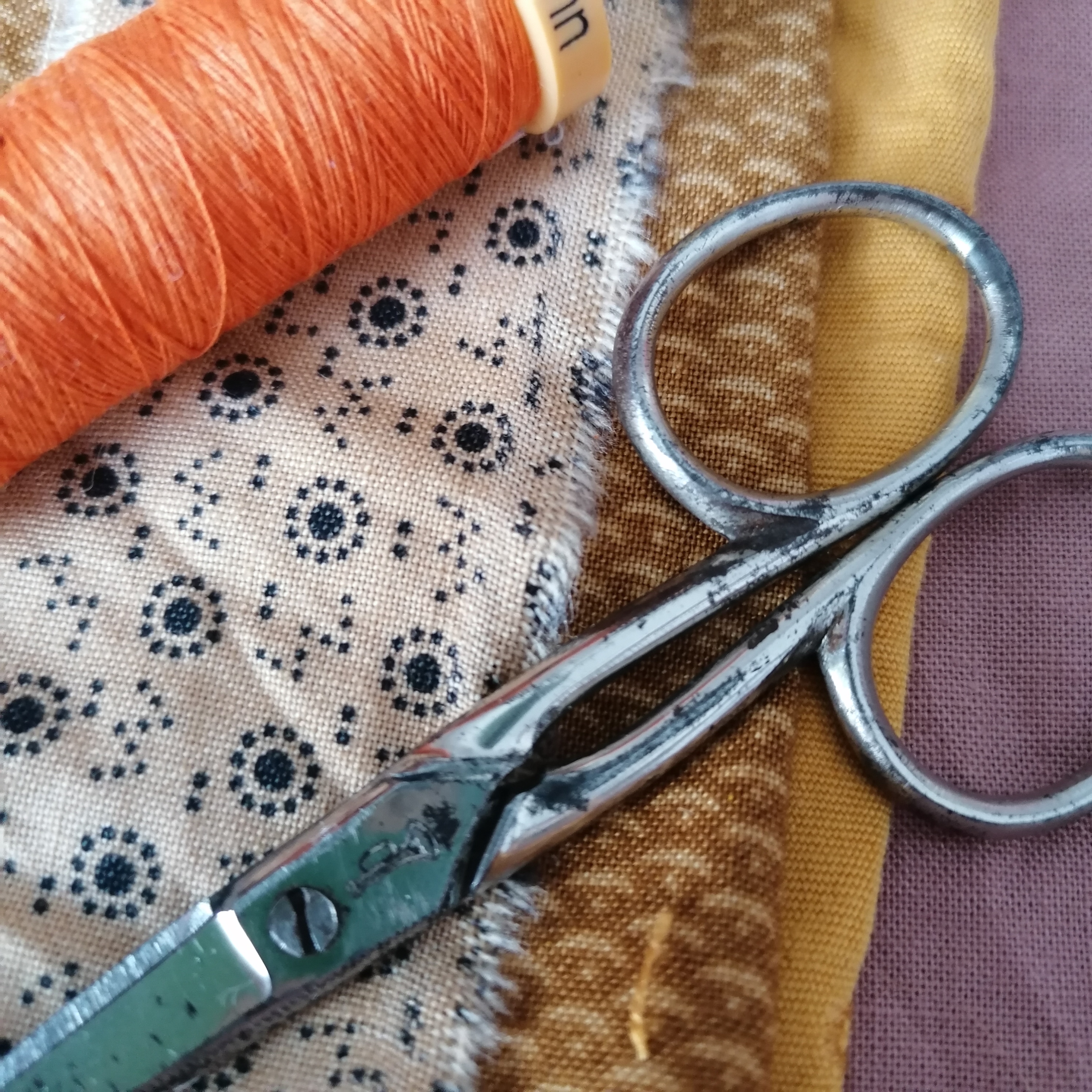 Stash of fabric in autumnal colours with orange thread bobbin and small metallic scissor