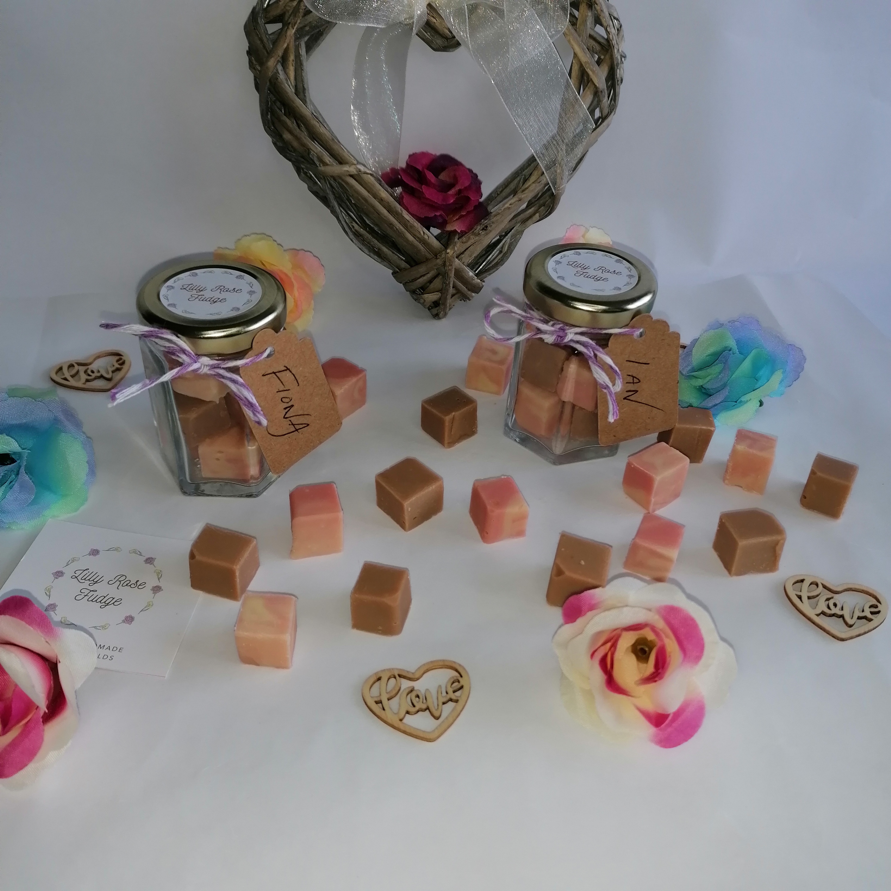 Two small glass jars filled with mini cubes of fudge, red and white twine with a brown kraft tag tied around the neck of the jar on the left and lilac and white twine with a kraft tag on the jar on the right. Cubes of fudge scattered around the bottom of the two jars. Behind the jars is a grey heart with a blue rose propped against the back.