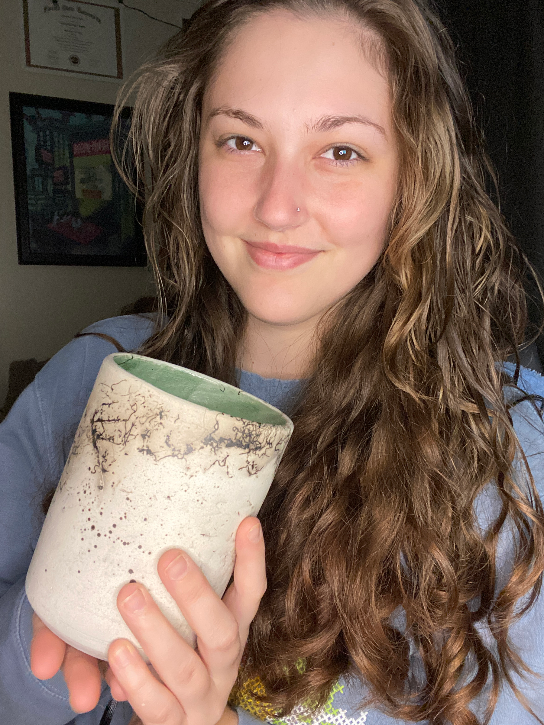 Emily is a young woman with long curly brown hair and brown eyes. She is wearing a blue sweatshirt and holding a cylindrical pot with black smokey dots and squiggly lines indicative of Raku firing.  