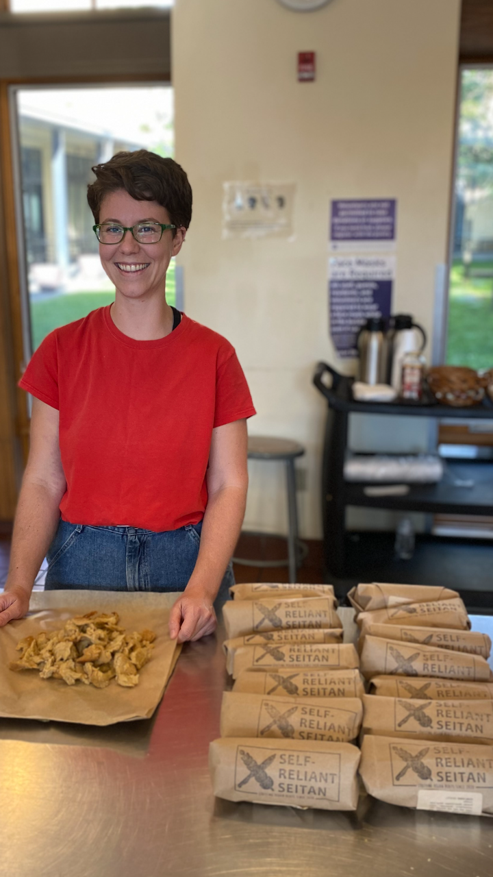 Image shows Emily Foster, the owner of Self-Reliant Seitan. She is a white woman with short light brown hair shown packaging seitan chickn shreds in brown paper packaging. Background is a commercial kitchen setting
