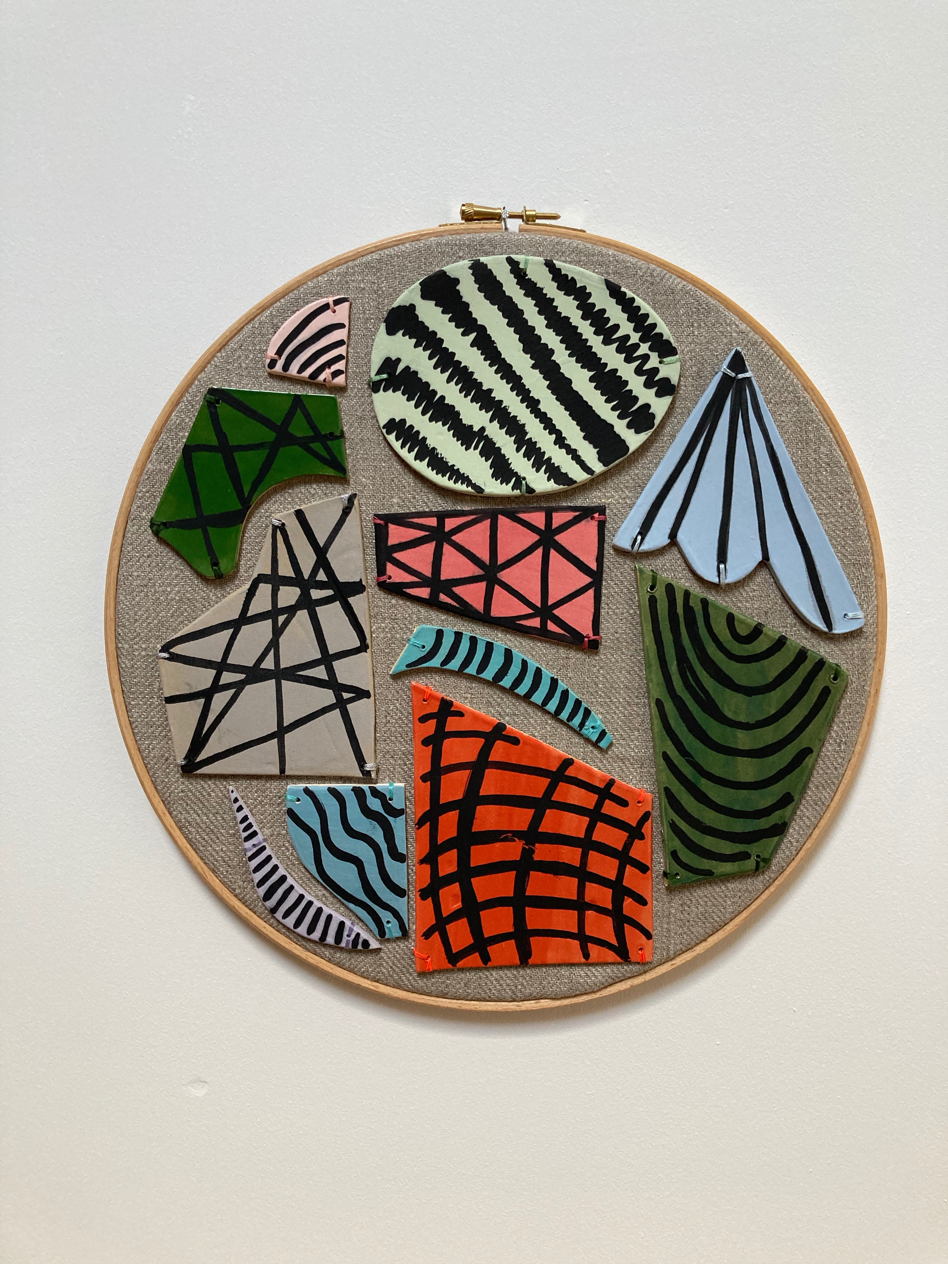 an embroidered hoop with pastel coloured ceramic shapes that have black ink painted marks and patterns 