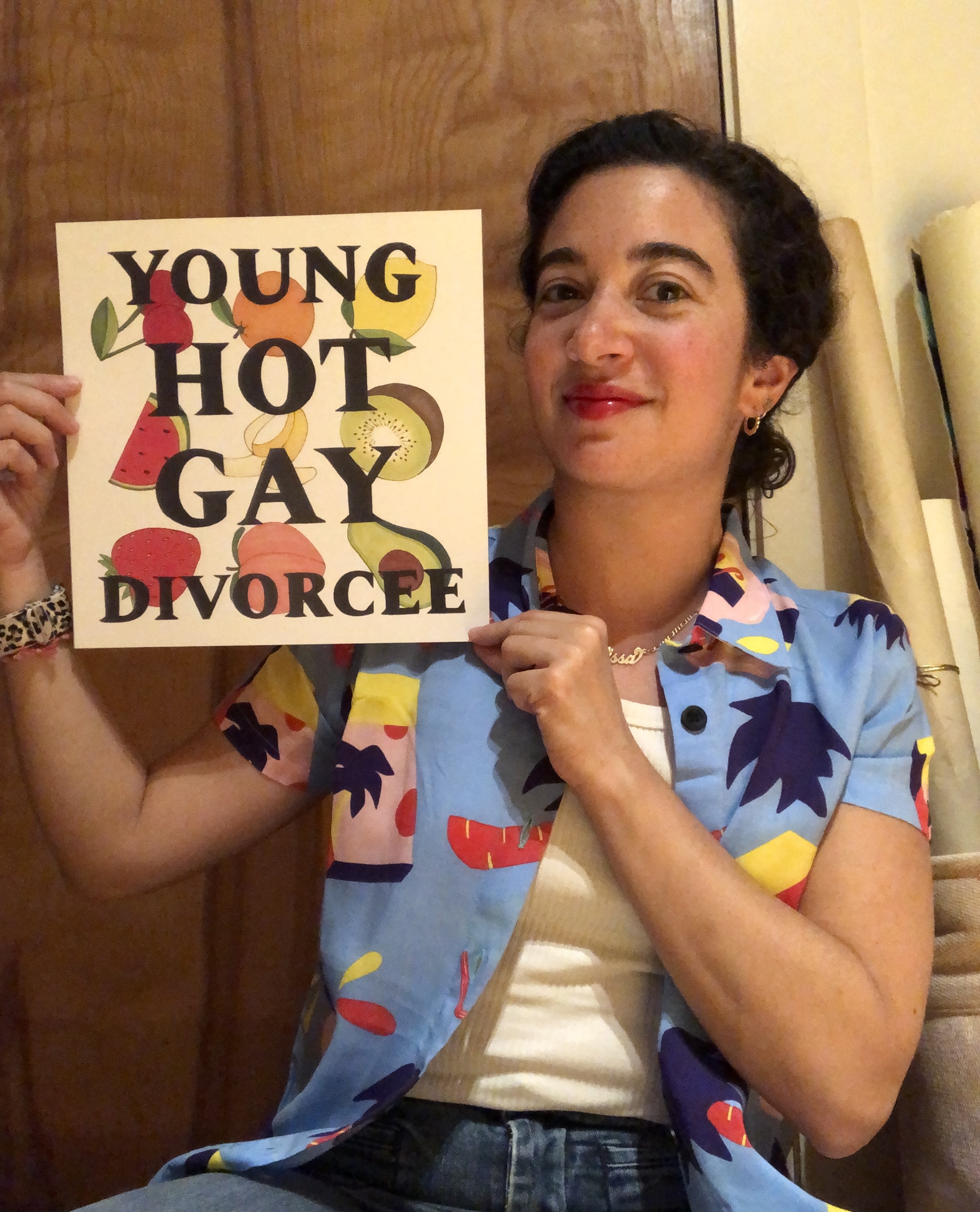 White femme with dark curly hair, wearing a colorful button-up shirt and holding a Young Hot Gay Divorcee print