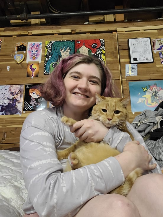 Femme presenting person holding an orange cat who does not appear happy about the picture at all