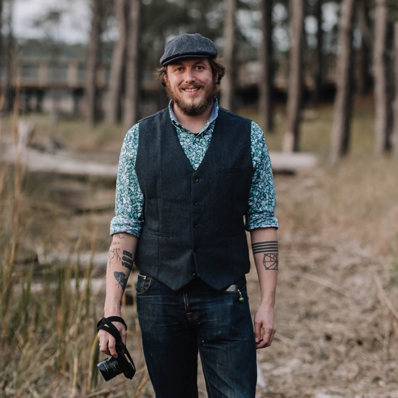 Jonathan stands near the water's edge, holding his camera, wearing a cap, herringbone brown vest, light green paisley shirt, and jeans. 
