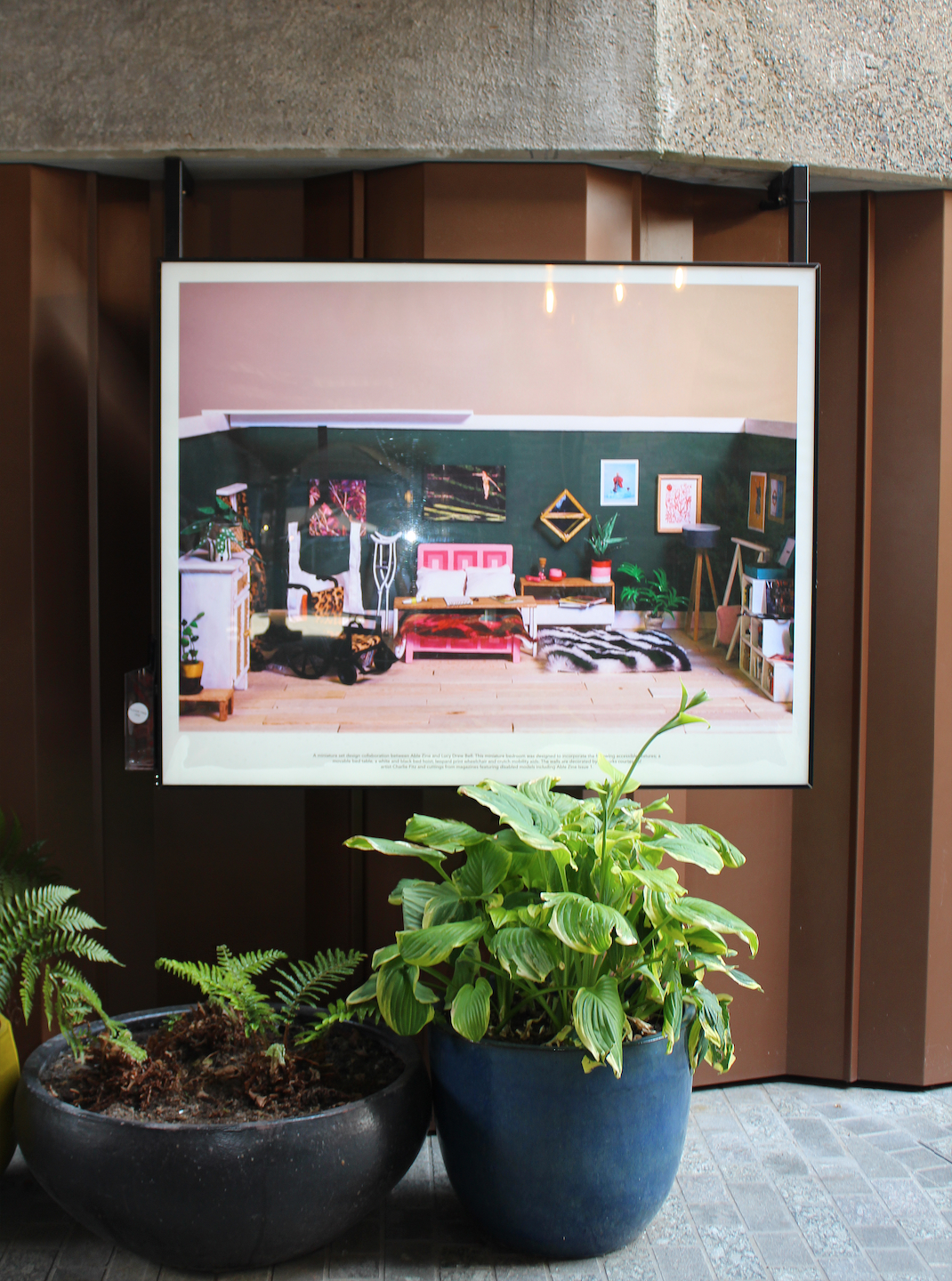 A photograph of a lightbox that shows a miniature model set of a bedroom. The bedroom is covered in unique decorations. It has a bed in the middle with a movable bed desk above, covered in art materials. There is also a leopard print wheelchair, crutches and a bed hoist in the room. On the floor is a fluffy zebra print rug. Outside of the lightbox theres a concrete wall and three trees/plants sat infront of the lightbox.