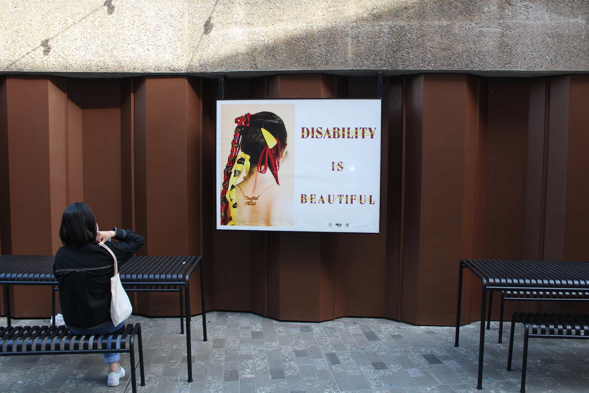 A woman is sat on a table facing away from the camera. In front of her in the centre is a lightbox containing two sides. On one side we see the back of a woman's head. Her hair is creatively parted revealing a large scar straight down her back.  Beneath the scar is a necklace that reads "Disabled & Proud." adjacent is typography reading “DISABILITY IS BEAUTIFUL” in red, whilst also reading “ACCESS IS BEAUTIFUL” in yellow
